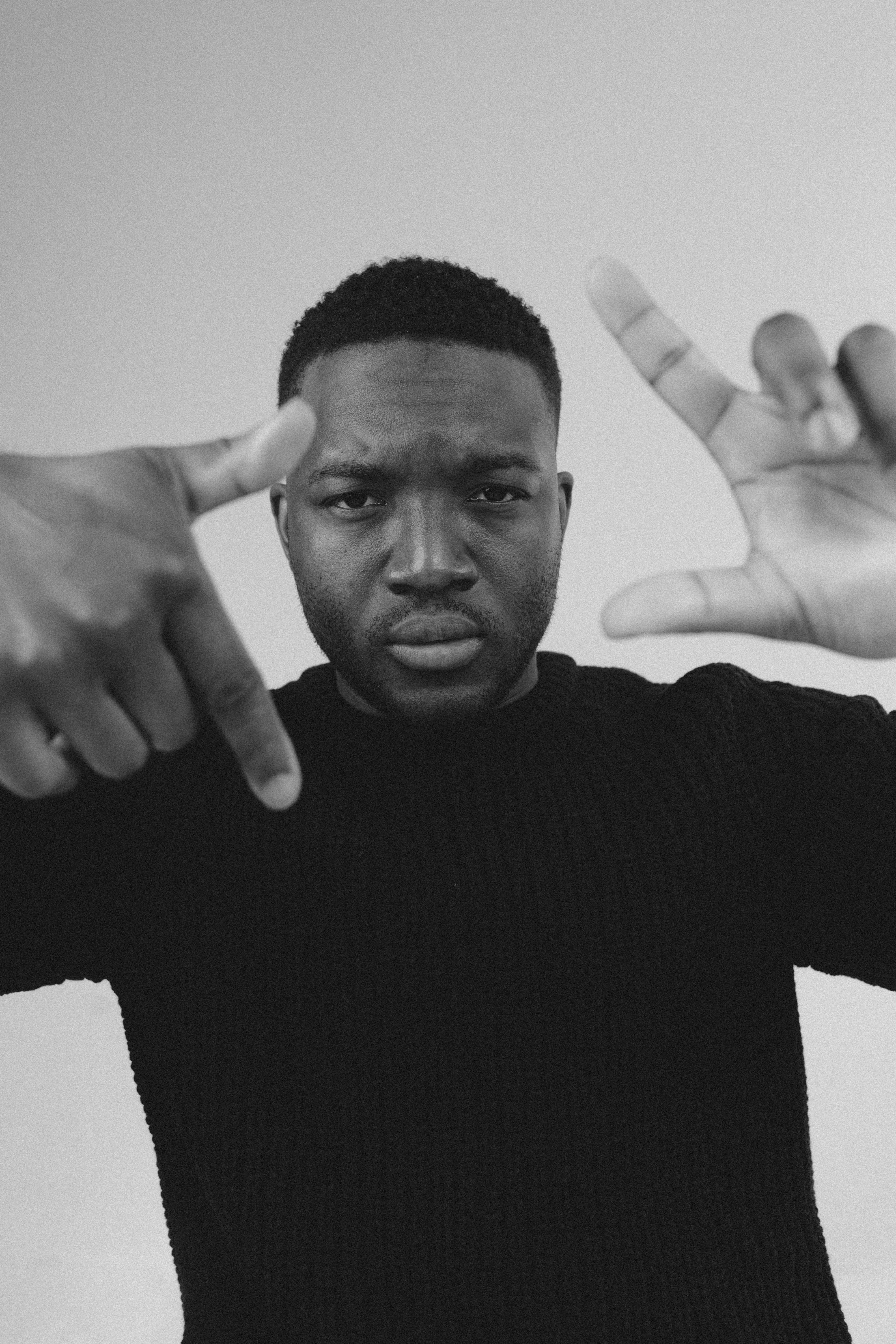 Young man with short hair making a gesture with both hands, pointing at his head and chest, against a plain background.