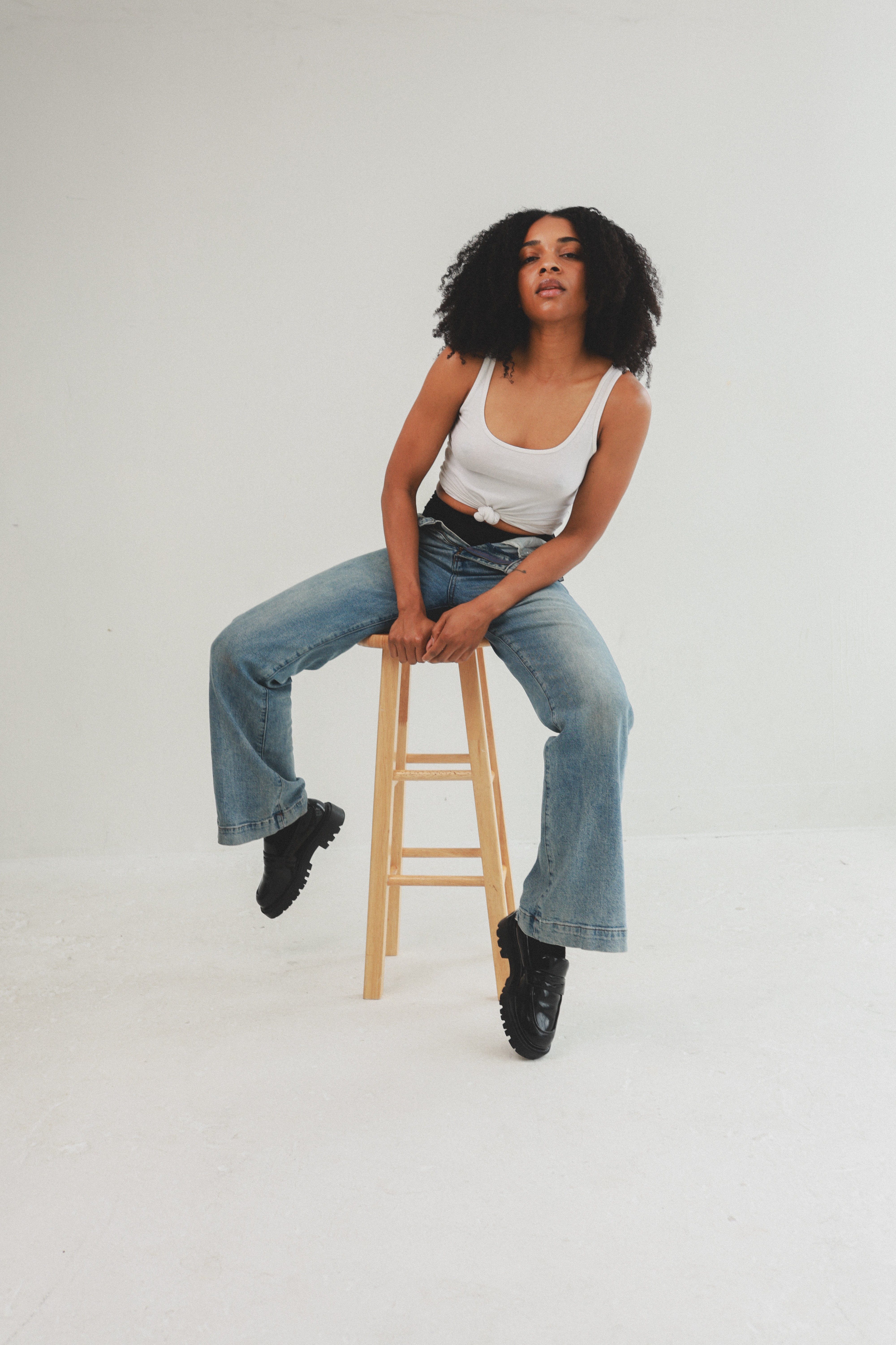 Woman with curly hair sitting on a wooden stool, wearing a white tank top and jeans, against a plain white background.