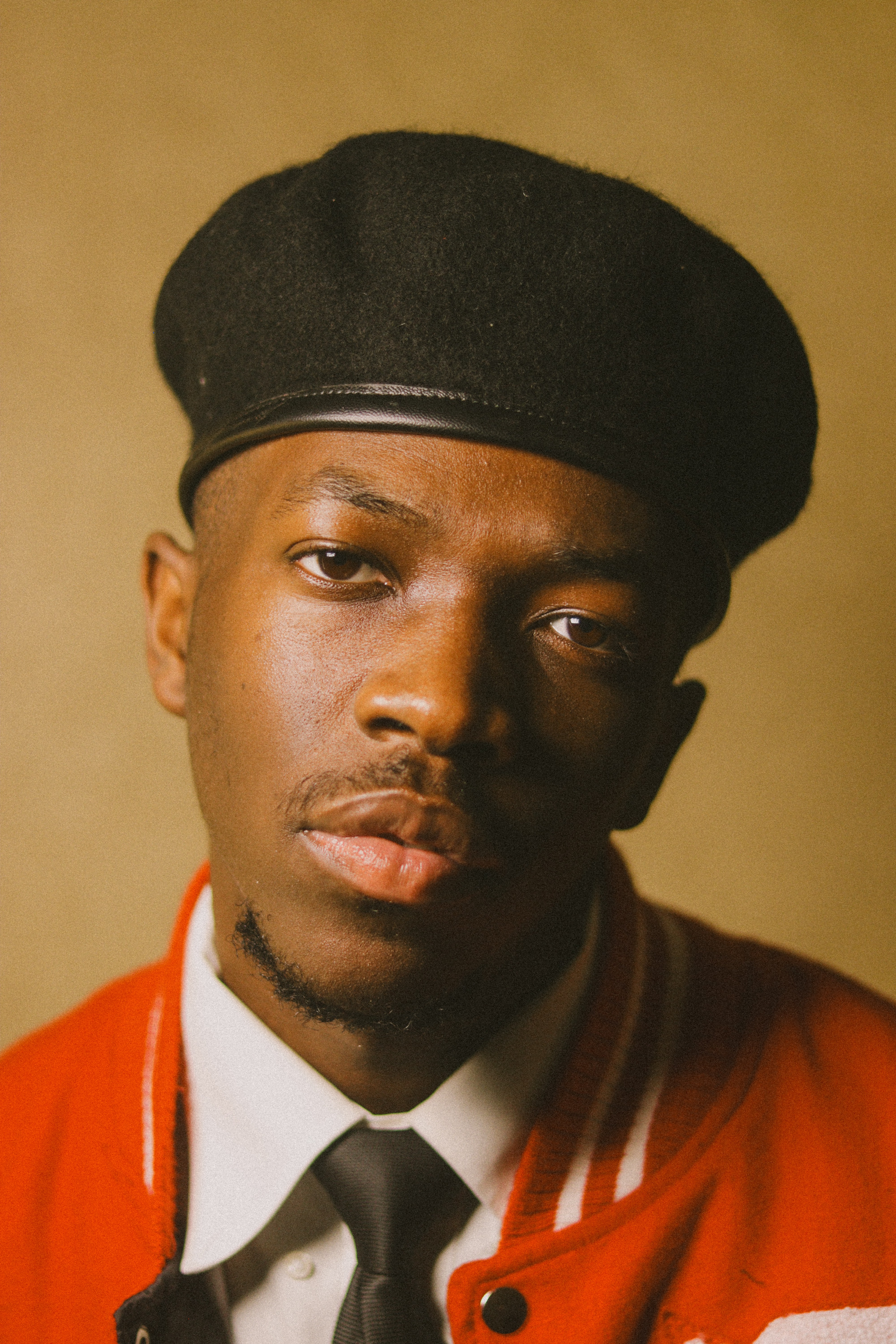 Young man wearing a black beret, red jacket, white shirt, and black tie, looking directly at the camera.