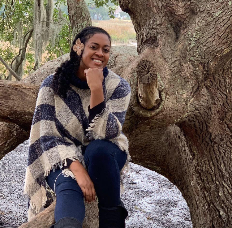 Woman sitting outdoors near a large tree, smiling, with a stream in the background.