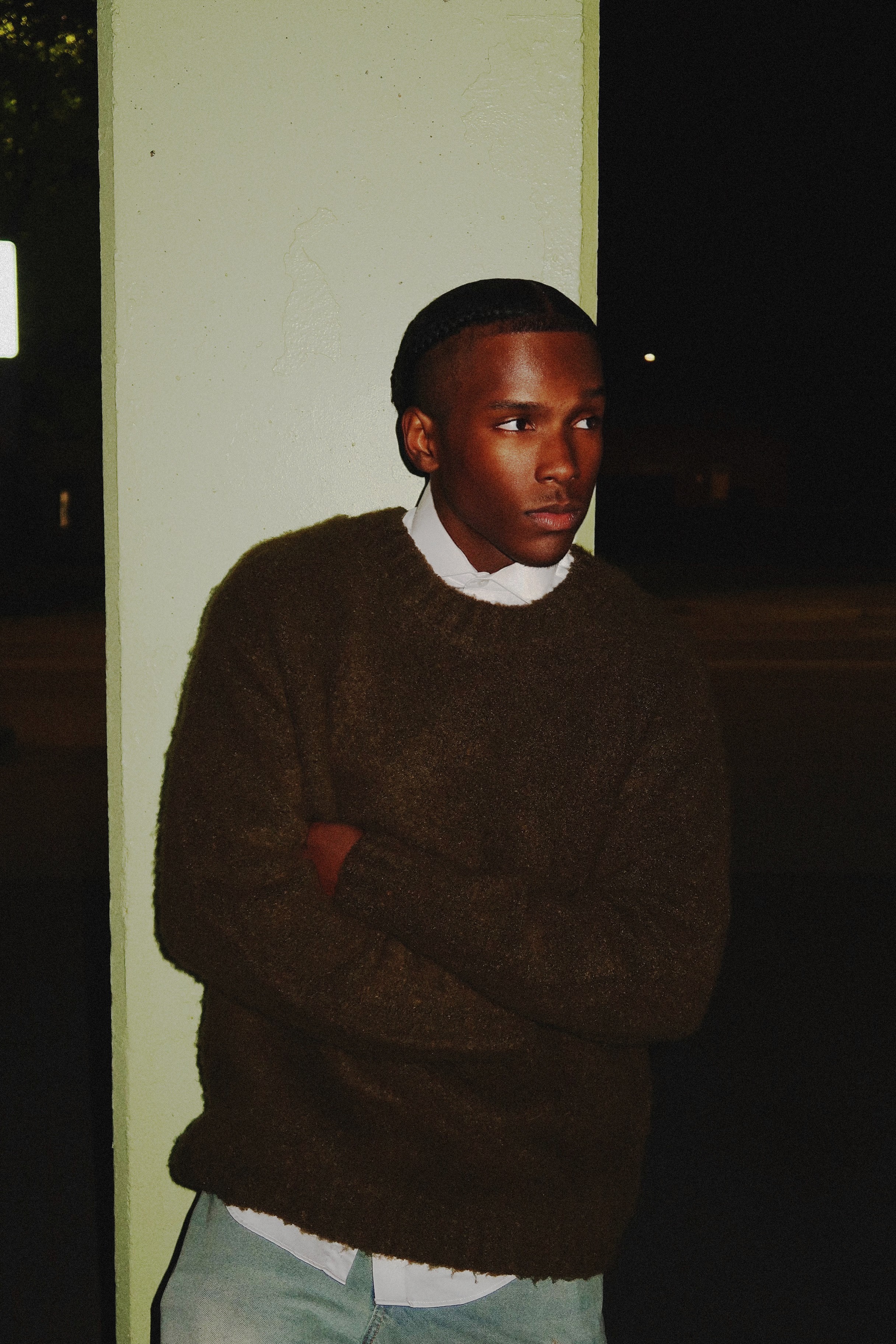 Young person with crossed arms, wearing a dark sweater and white shirt, standing indoors near a light-colored wall.