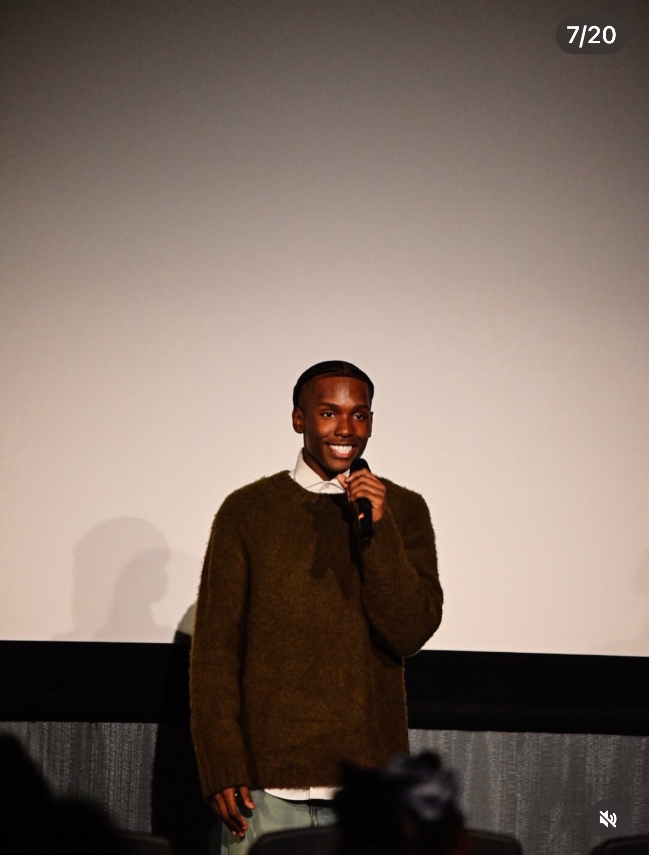 Person standing on stage holding a microphone, smiling, with a blank screen behind them.
