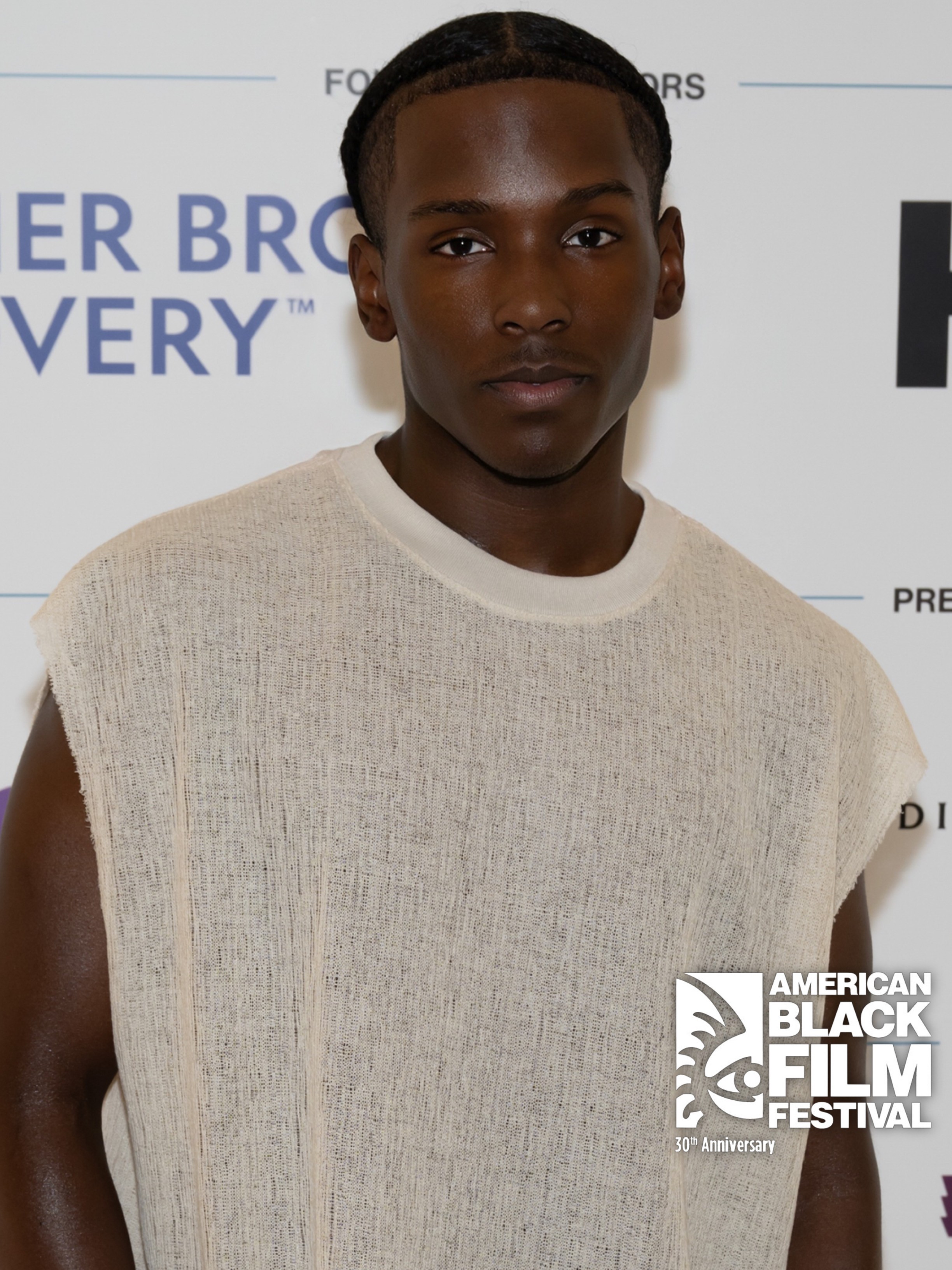 Young man with dark skin and braided hair wearing a sleeveless beige top at an event.