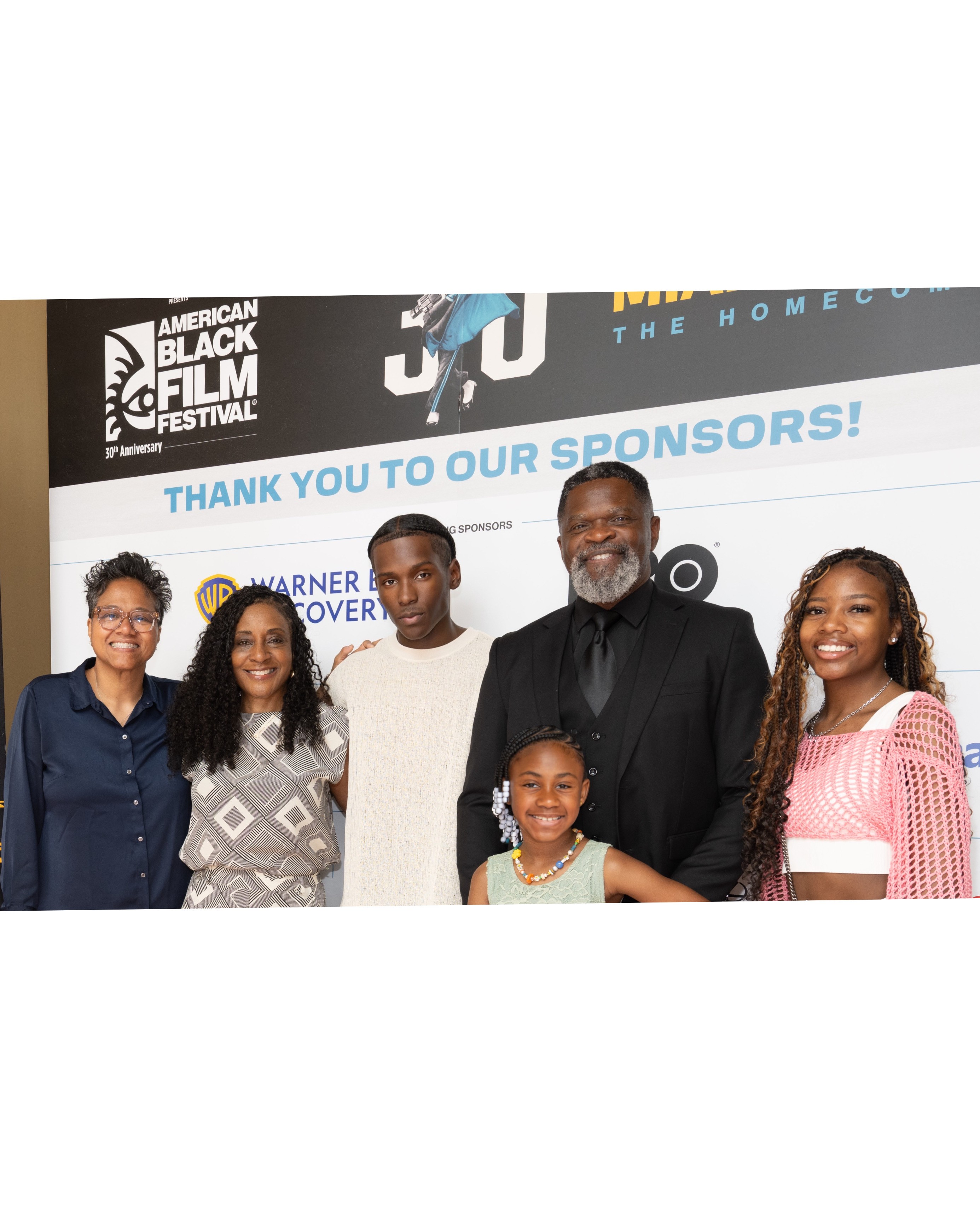 Group of six diverse people standing in front of a banner, smiling, with text thanking sponsors, indoors.