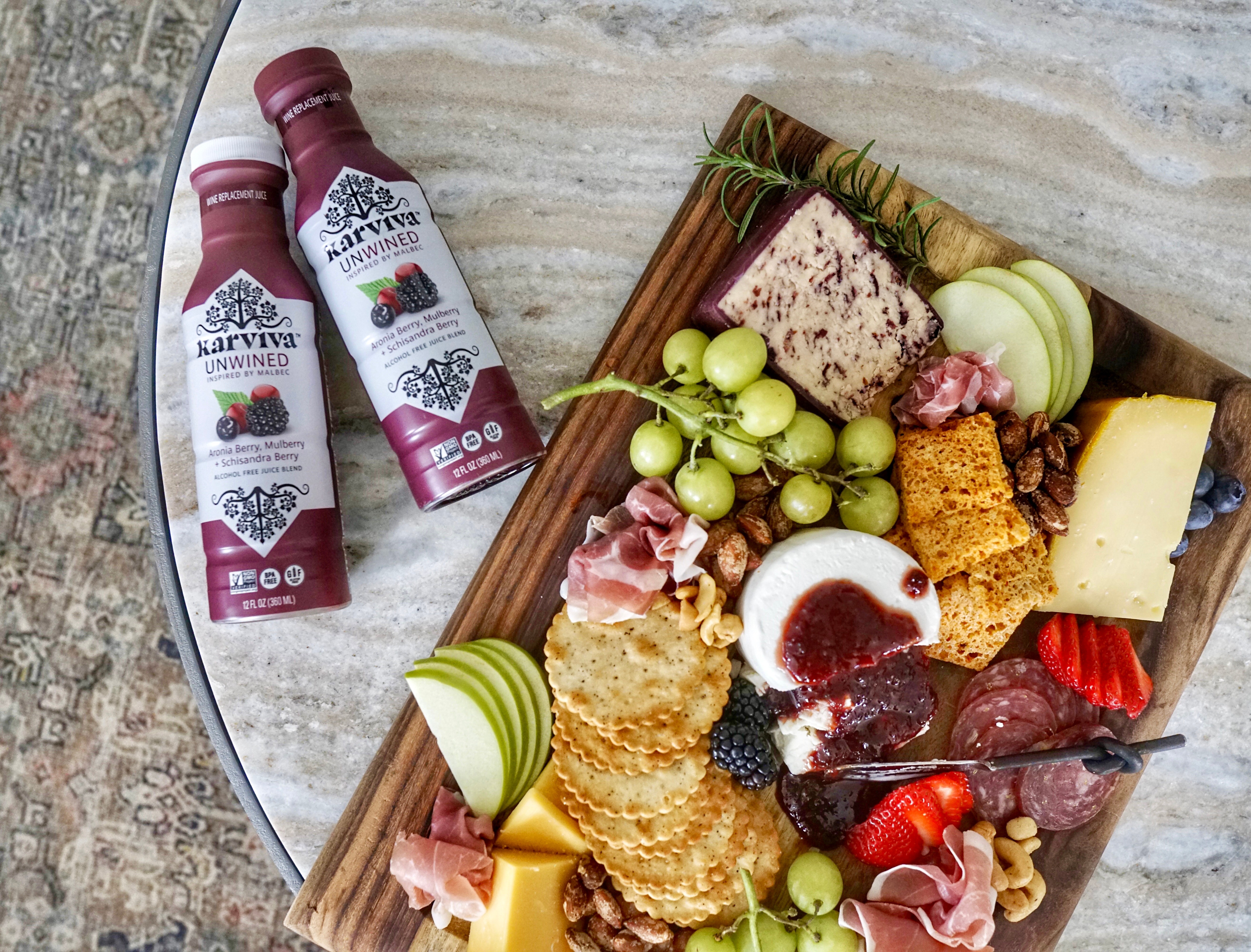 A wooden tray with cheese, grapes, strawberries, bread, and spreads on a marble surface.