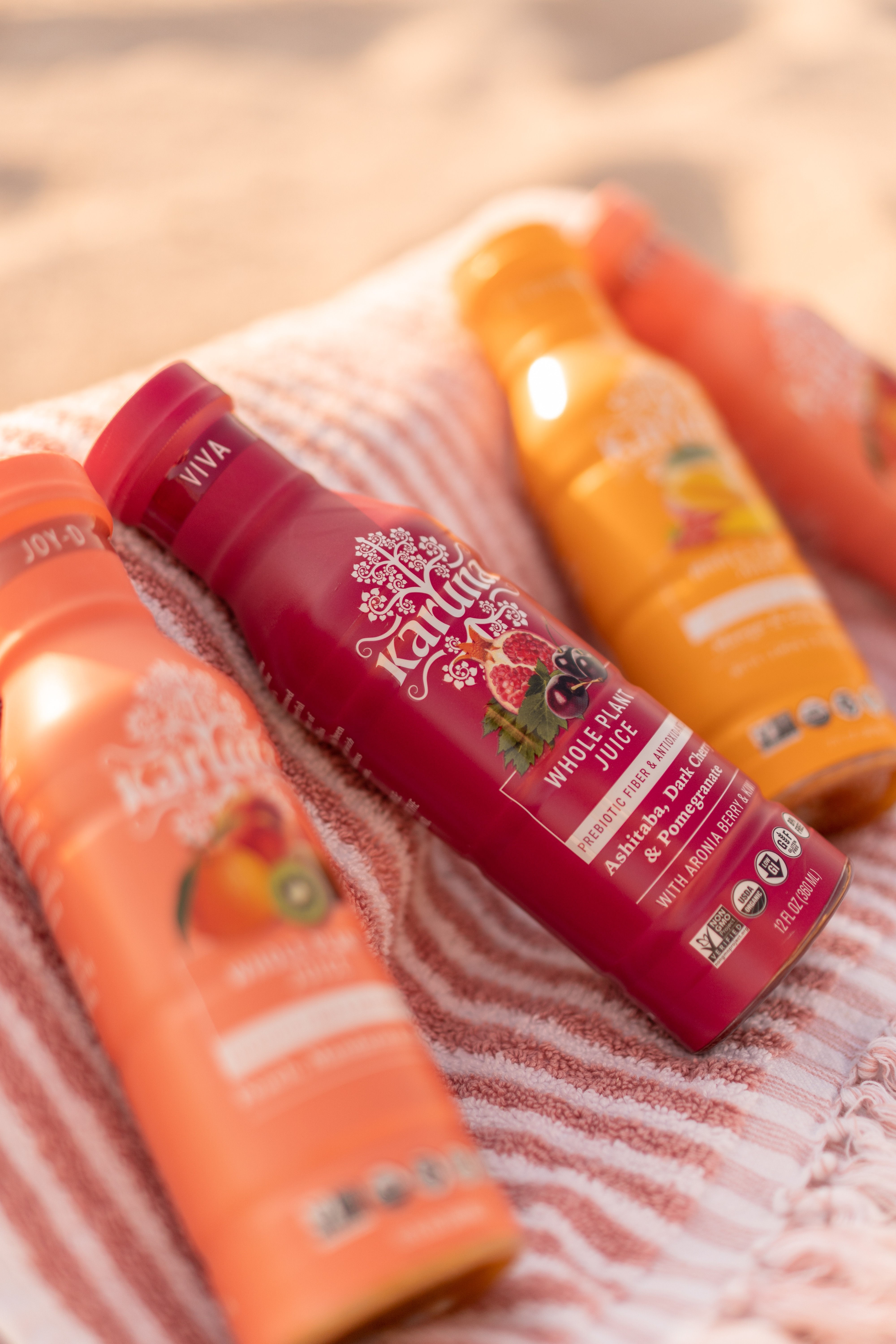 Four bottles of fruit-flavored drinks on a striped towel, with a blurred sandy background.