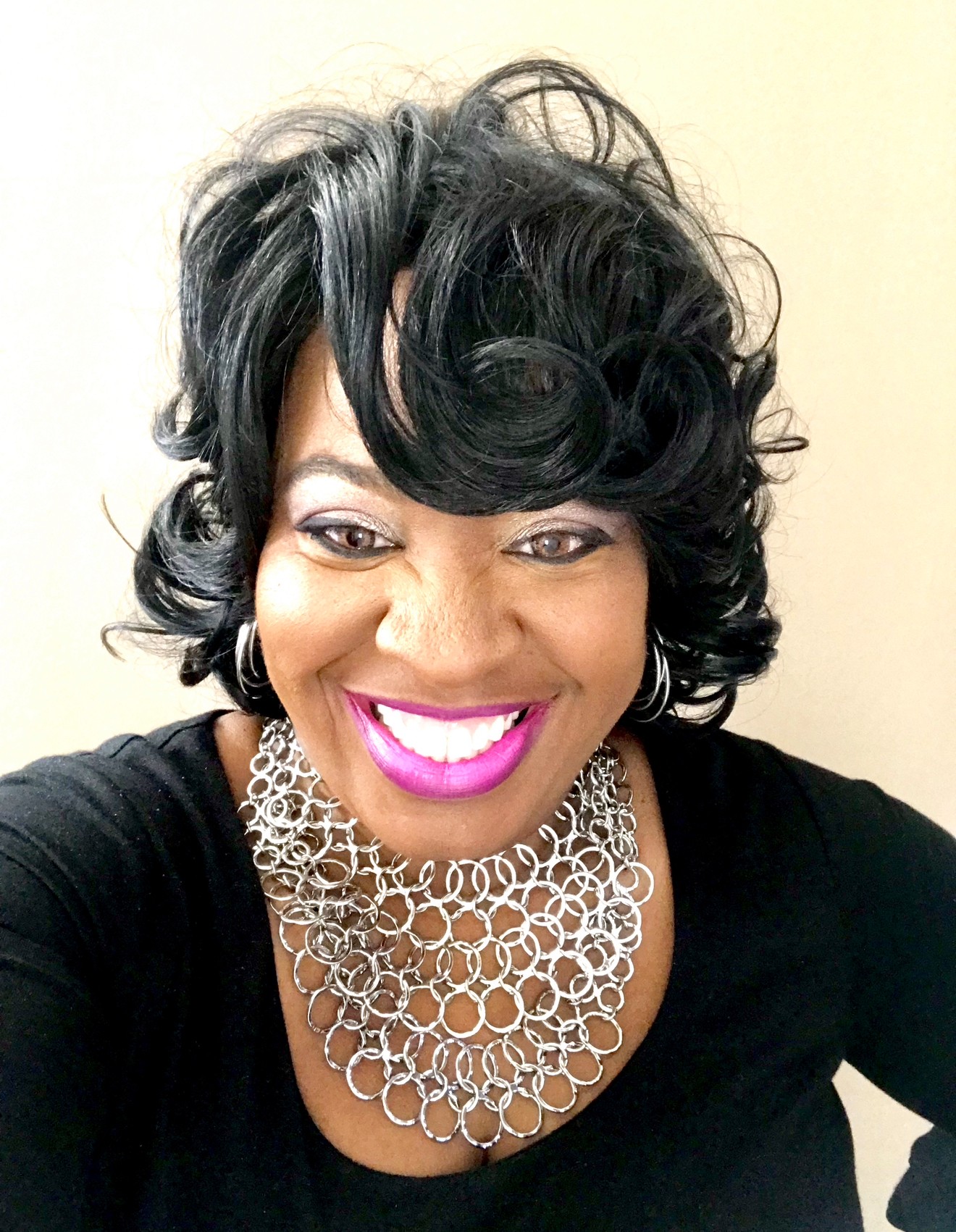 Woman with short, curly black hair, smiling, wearing a black top and a large silver necklace.