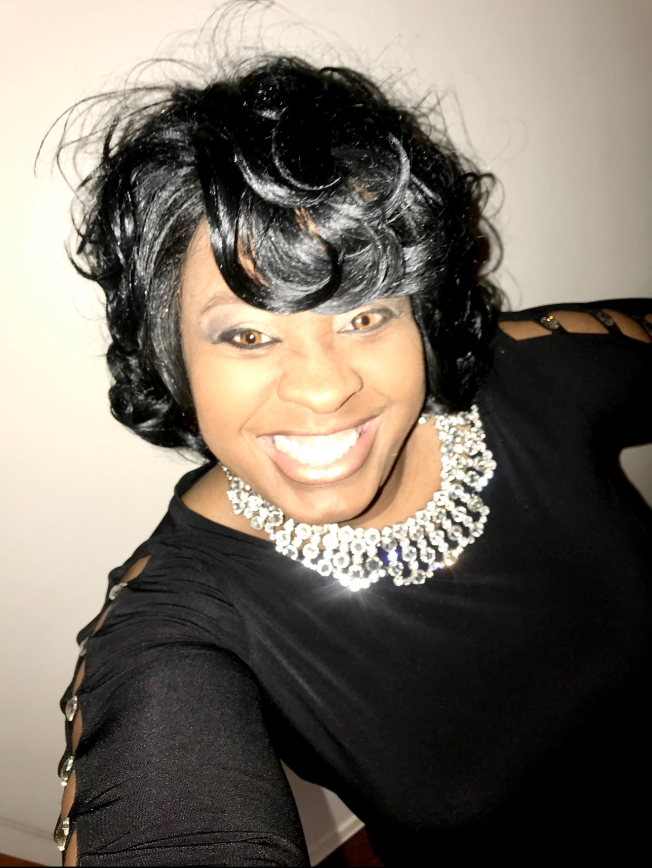 Woman with black curly hair smiling, wearing a black top and a pearl necklace, against a plain background.