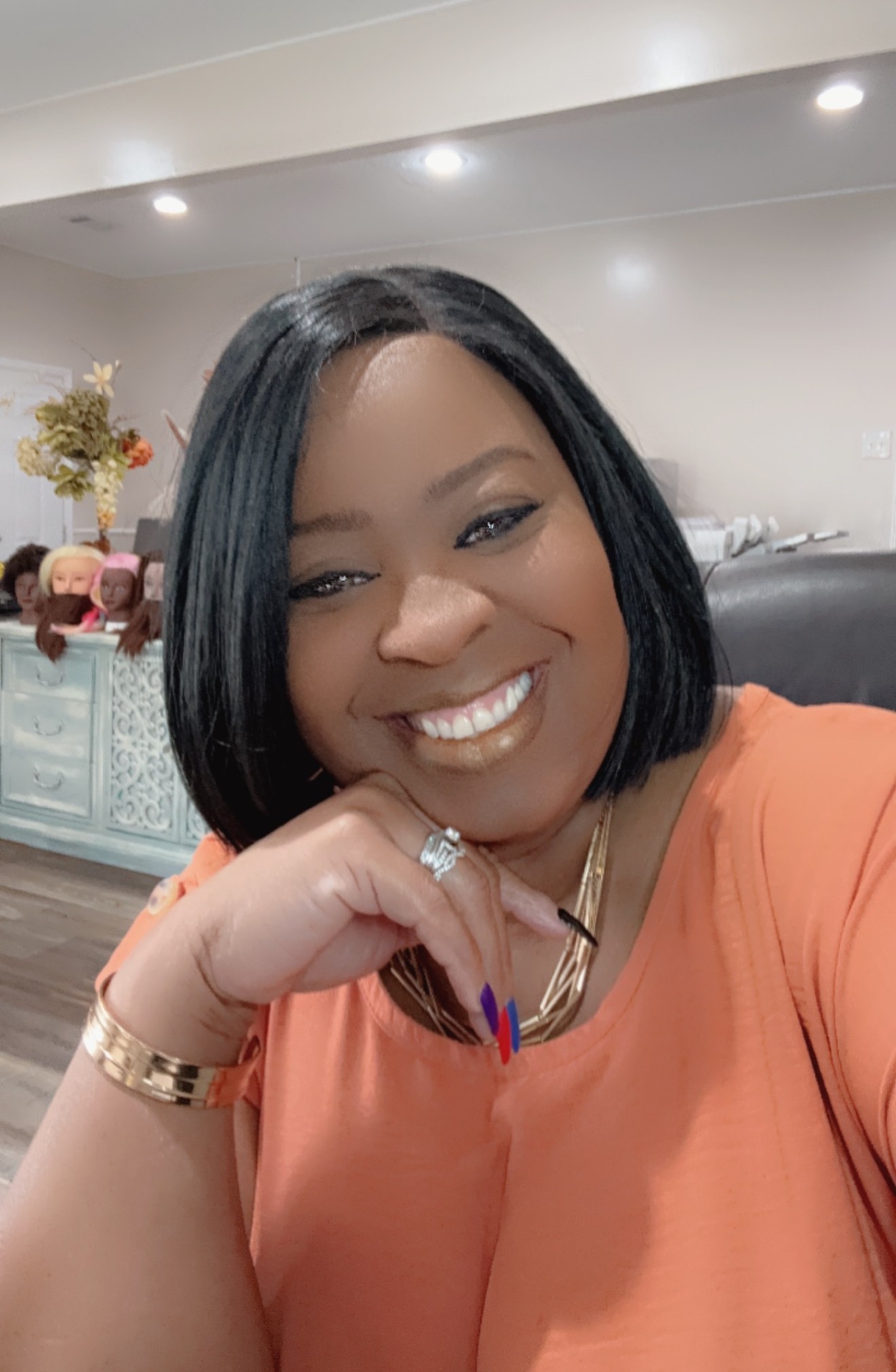 Smiling woman with shoulder-length black hair, wearing an orange top, jewelry, and sitting indoors.