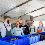 Five people stand behind a counter with blue containers, smiling and talking inside a metal building.