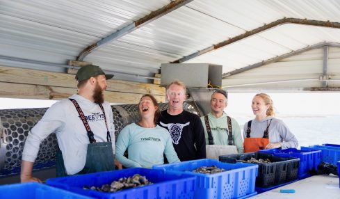 Five people stand behind a counter with blue containers, smiling and talking inside a metal building.