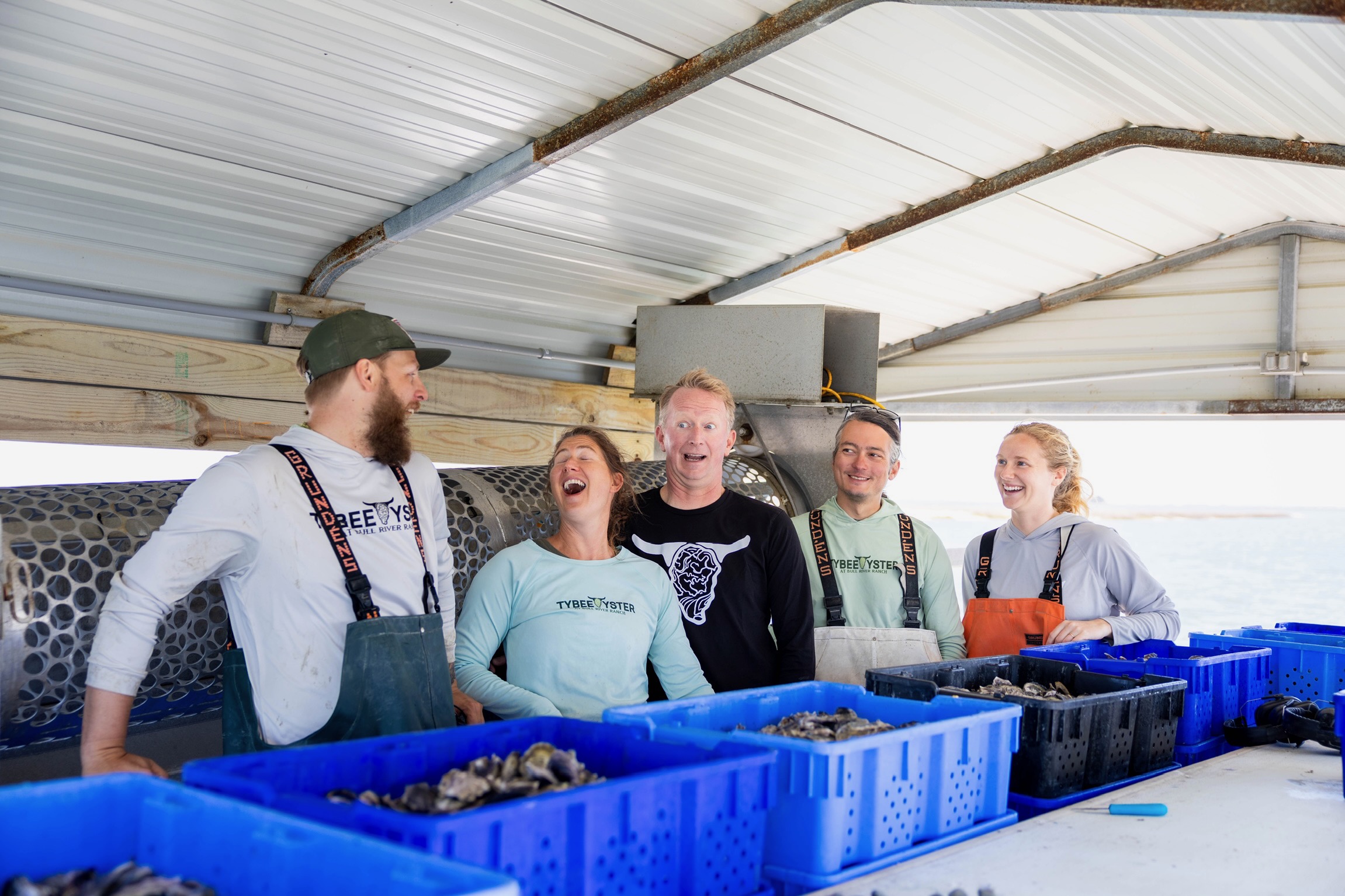 Five people stand behind a counter with blue containers, smiling and talking inside a metal building.