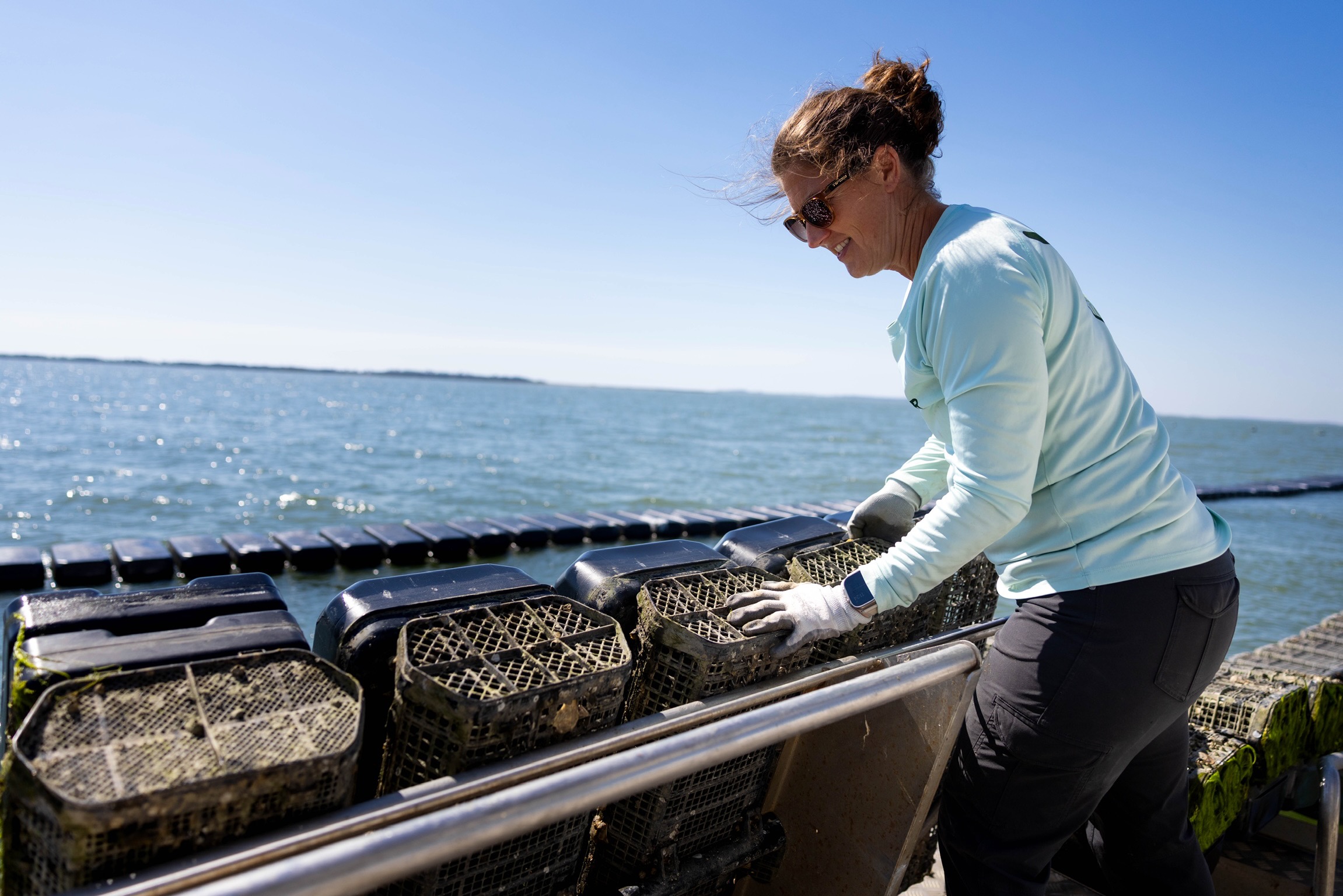 Woman wearing sunglasses and gloves, leaning over a railing by the water, handling lobster traps on a sunny day.