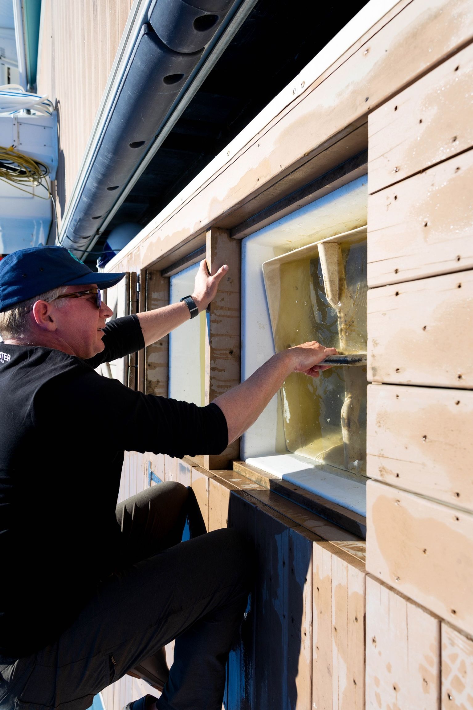 Person kneeling on a wooden dock, reaching into a glass-bottom boat window, with water visible inside. Word count: 20.