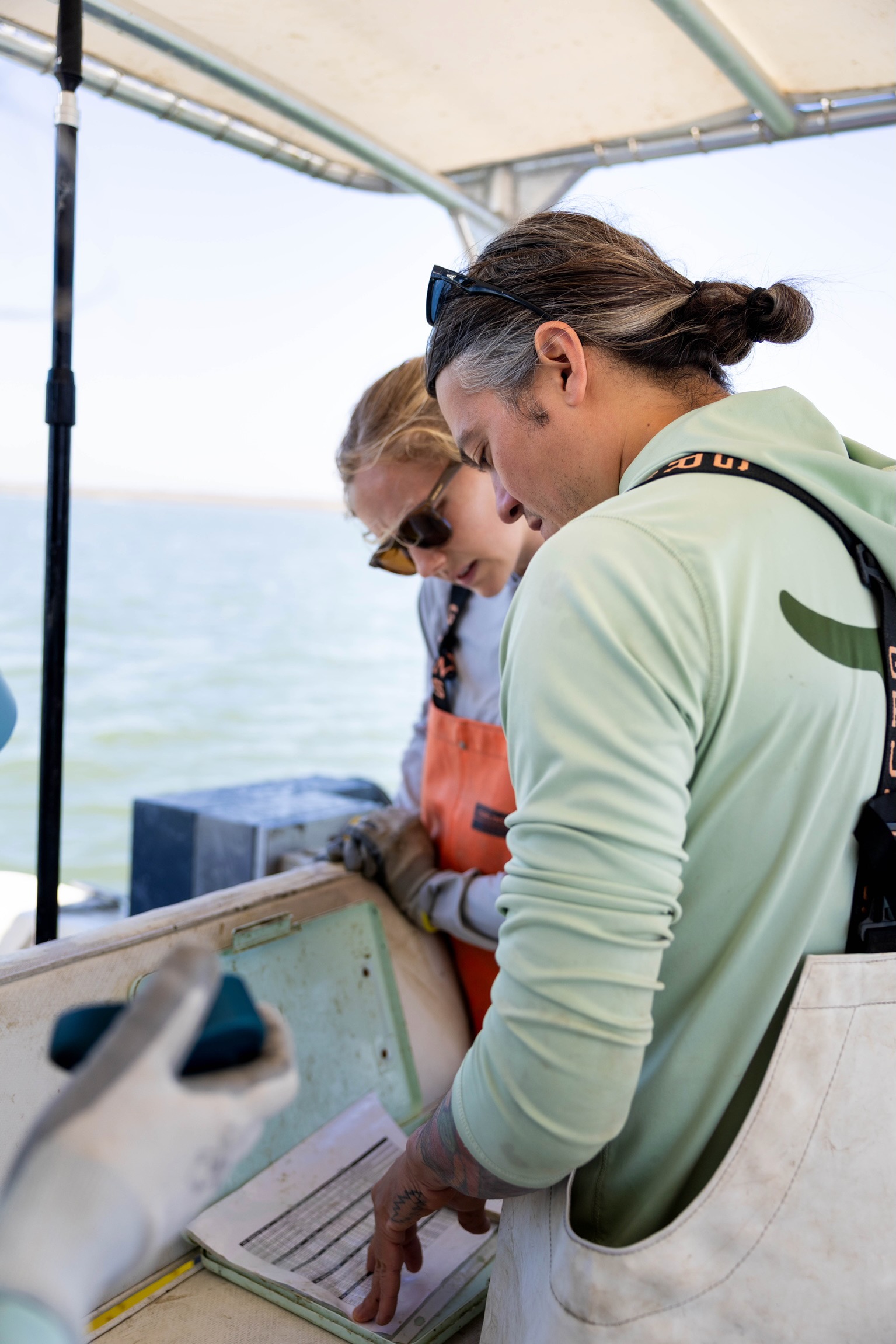 Two women on a boat looking at a clipboard, with water in the background, under a canopy.