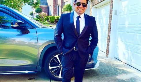Man in a suit standing outside near a car and garage, smiling, with houses and trees in the background.