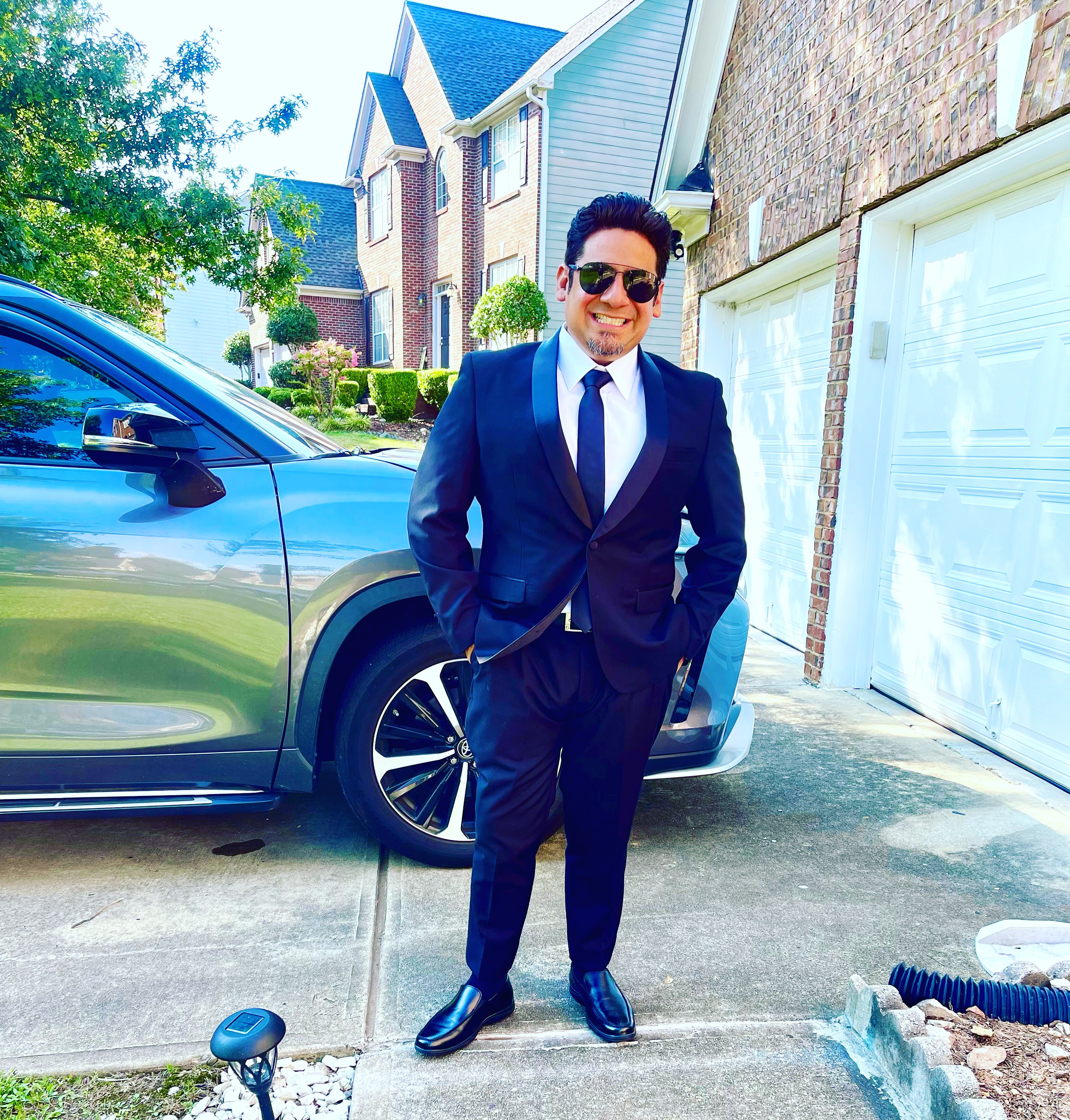 Man in a suit standing outside near a car and garage, smiling, with houses and trees in the background.