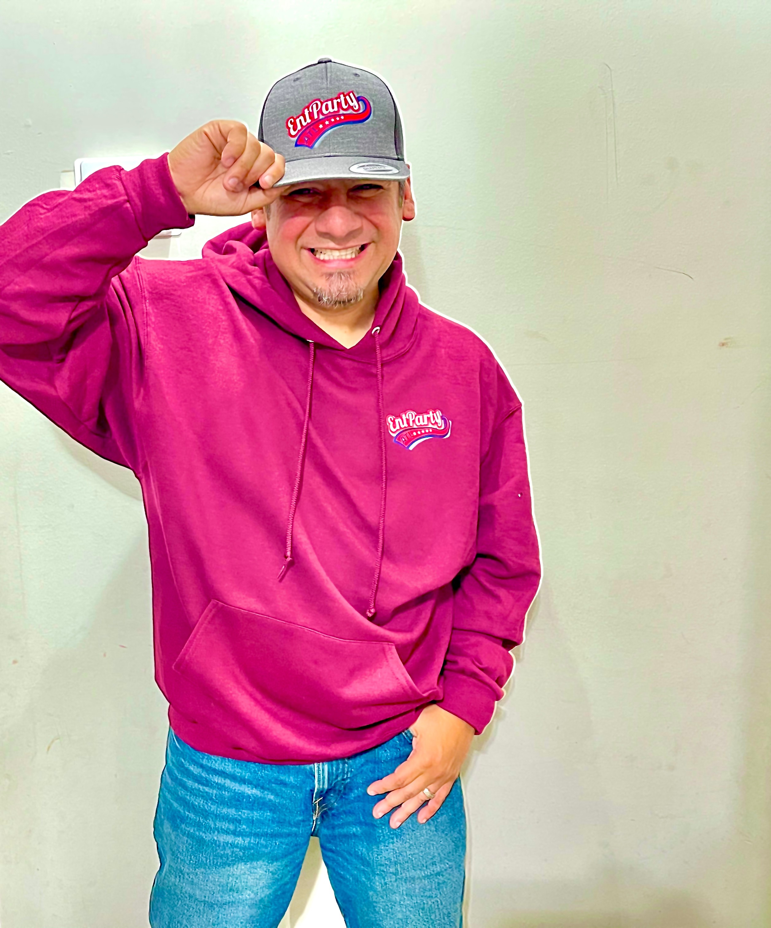 Smiling man in pink hoodie and gray cap, holding the cap's brim, standing against a plain background.
