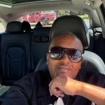 Person wearing sunglasses sitting in car with sunroof, looking at camera, hand near chin, parked outdoors.