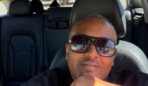 Person wearing sunglasses sitting in car with sunroof, looking at camera, hand near chin, parked outdoors.