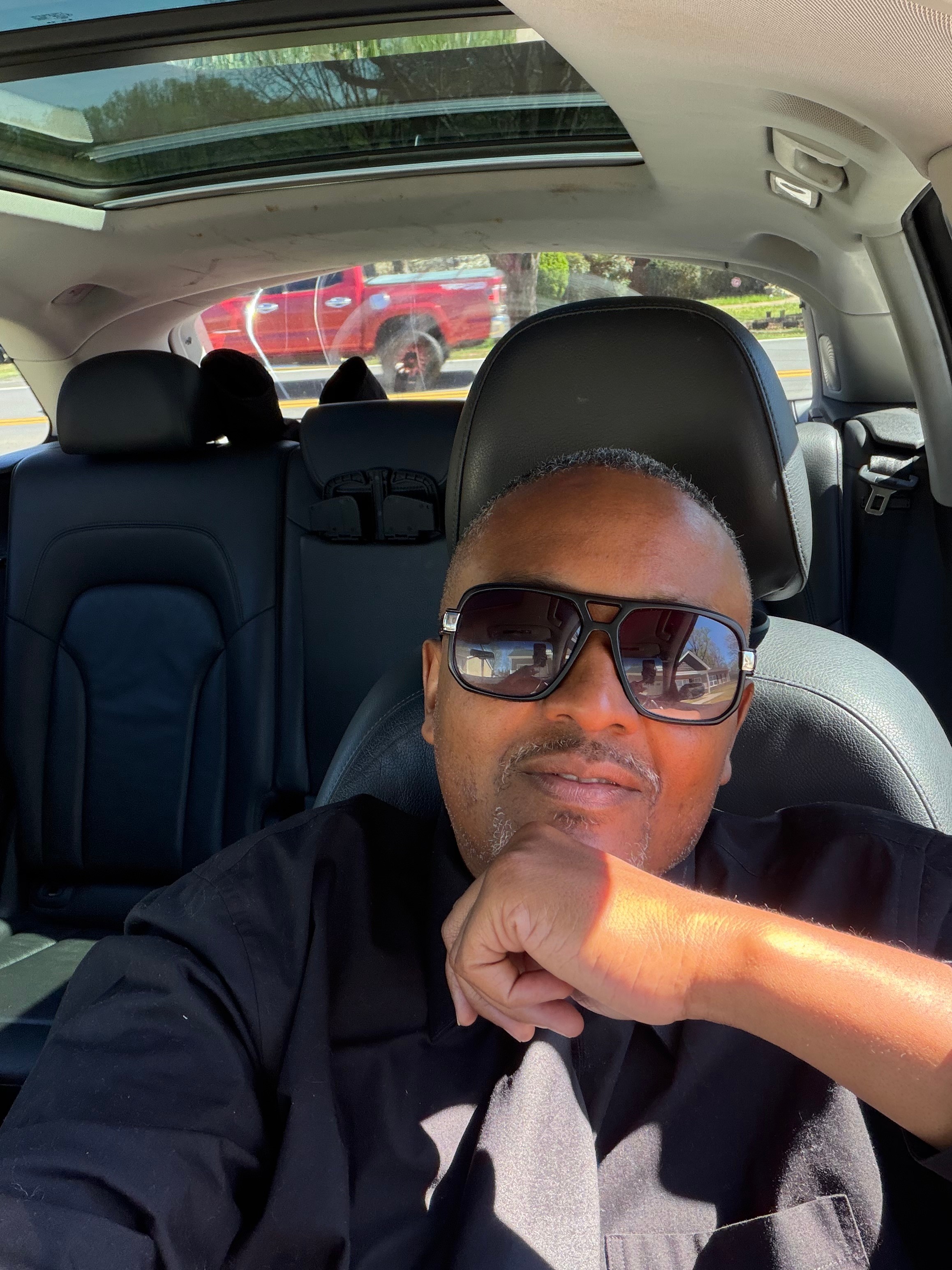 Person wearing sunglasses sitting in car with sunroof, looking at camera, hand near chin, parked outdoors.