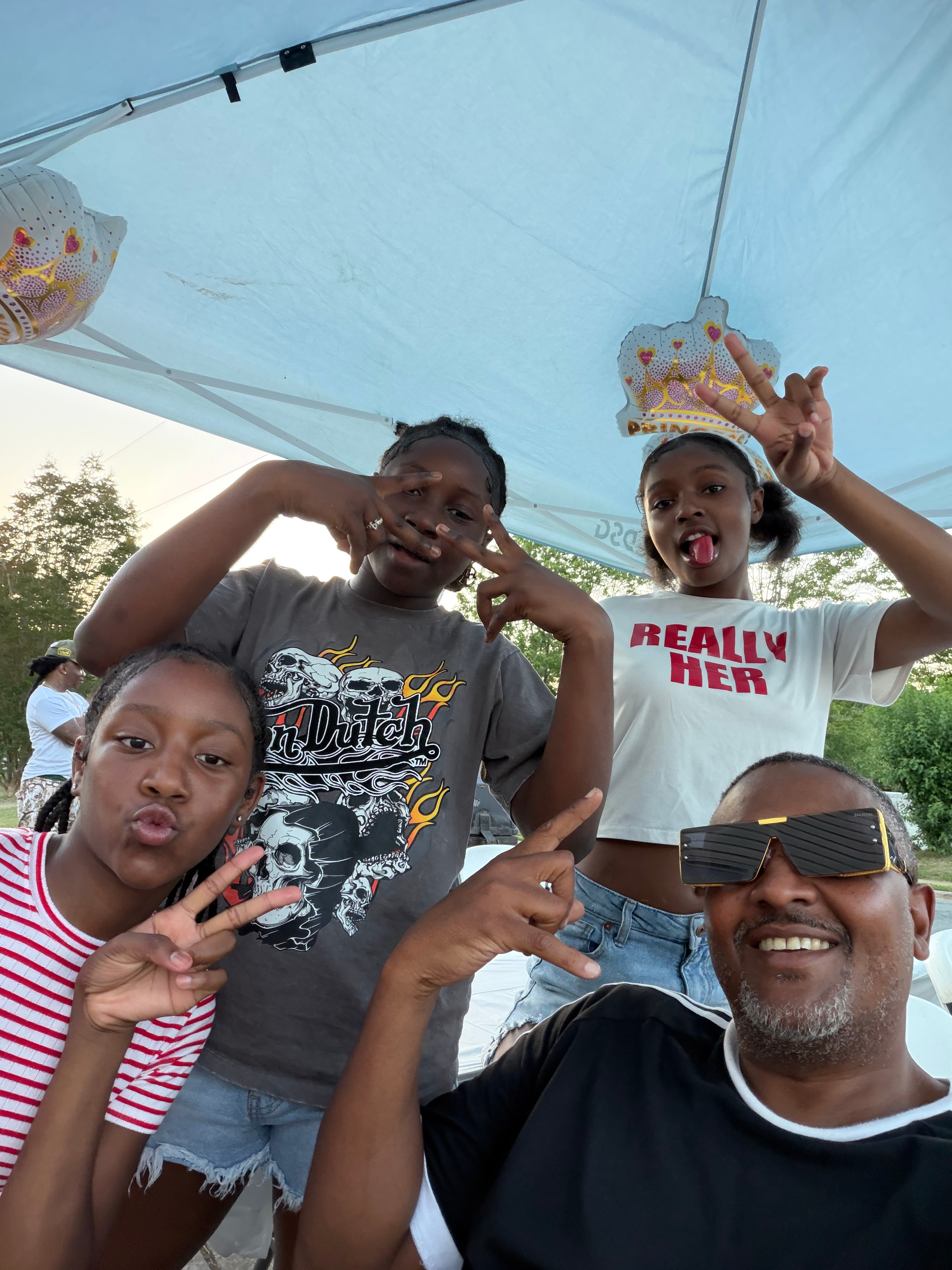 Four people under a blue canopy, two children and two adults, smiling and making peace signs, celebrating a birthday with crowns.