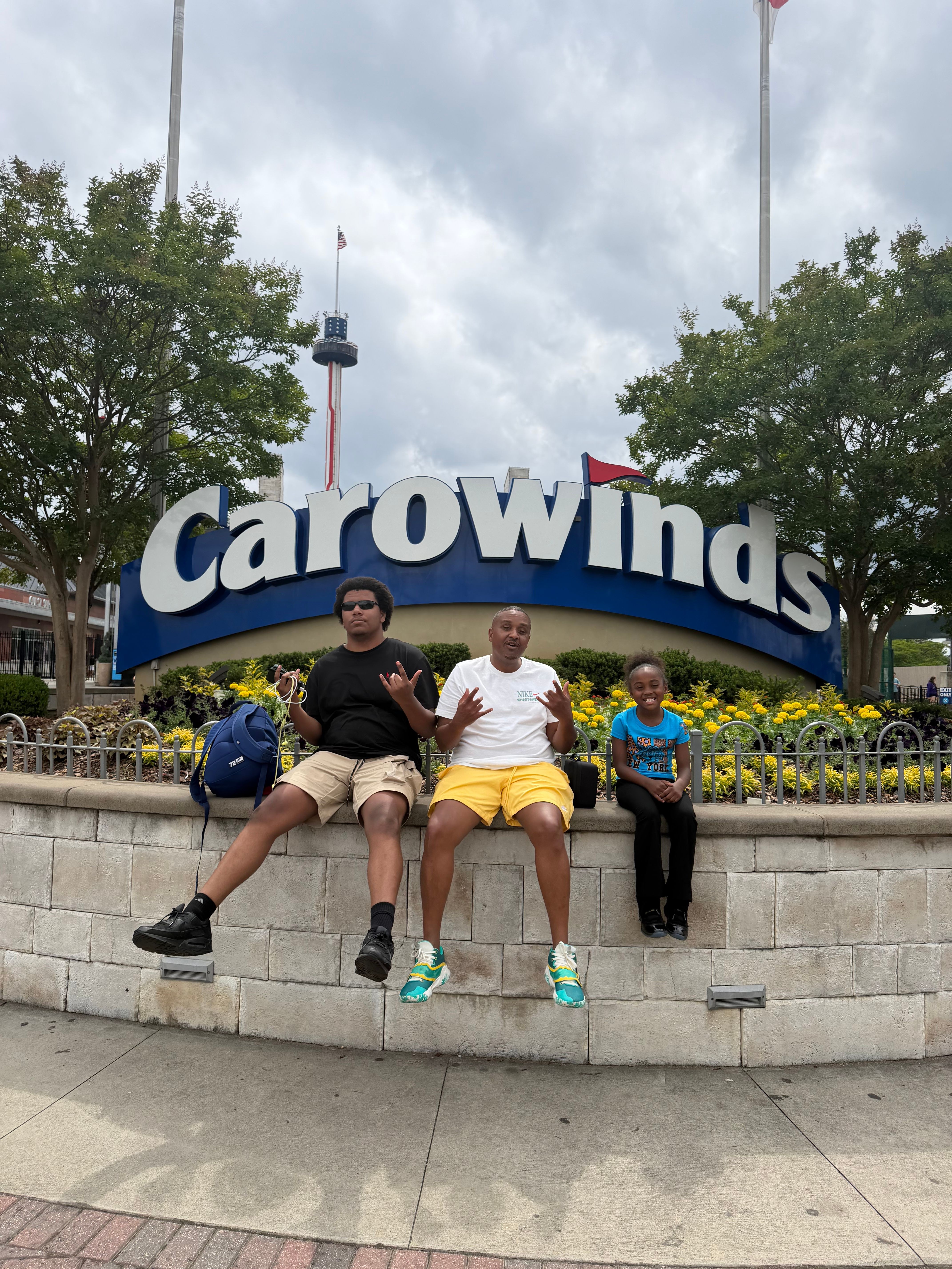 Two people sitting on a bench in front of a Caloundra sign, with trees and flowers nearby, under cloudy sky.