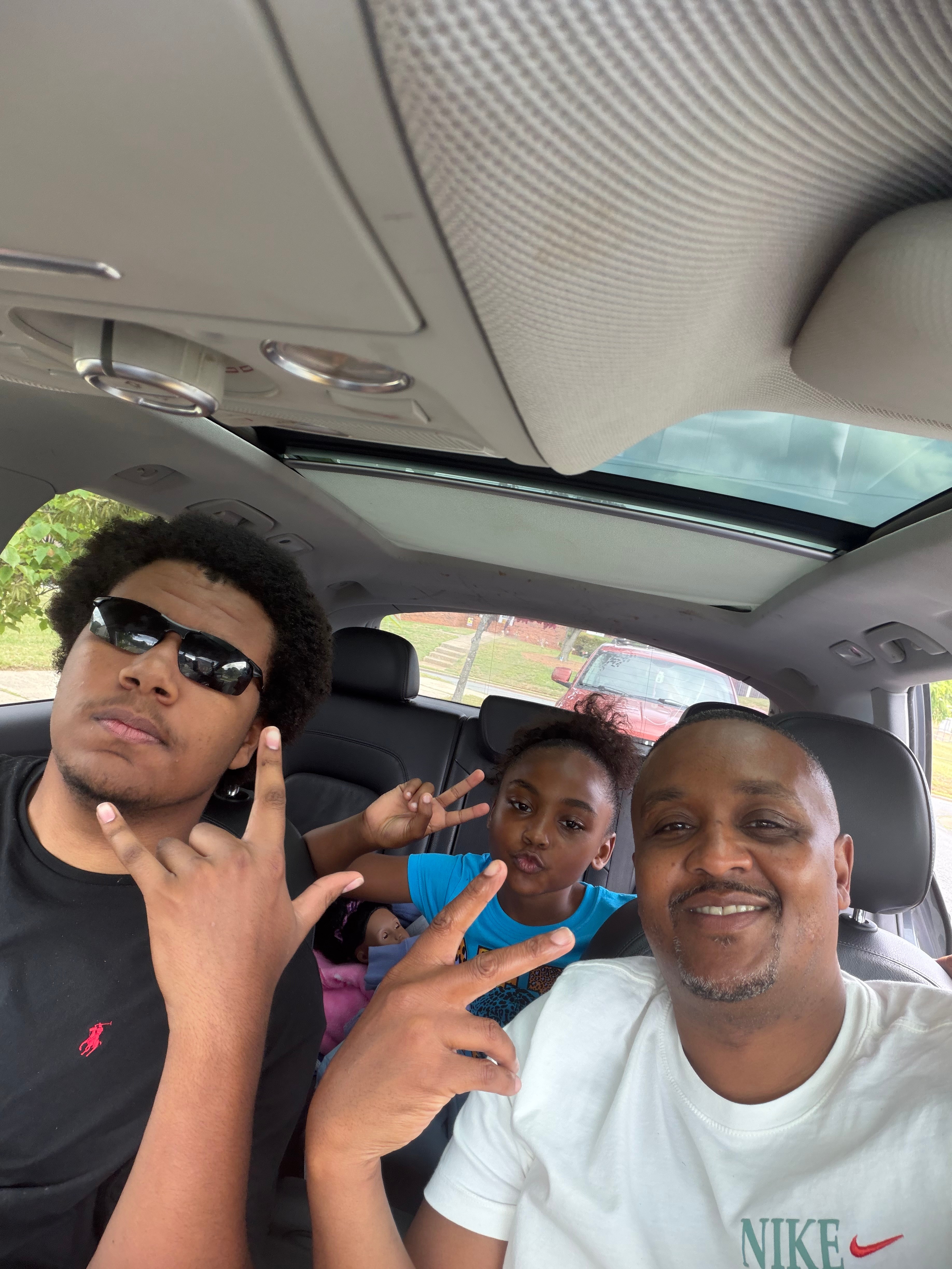 Three people sitting in a car, smiling and making peace signs, with trees and cars visible outside the window.