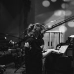 Person with curly hair working on a laptop in a dimly lit room with stage lights and equipment.