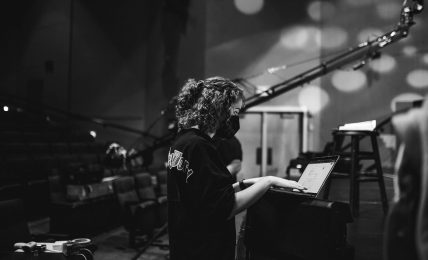 Person with curly hair working on a laptop in a dimly lit room with stage lights and equipment.