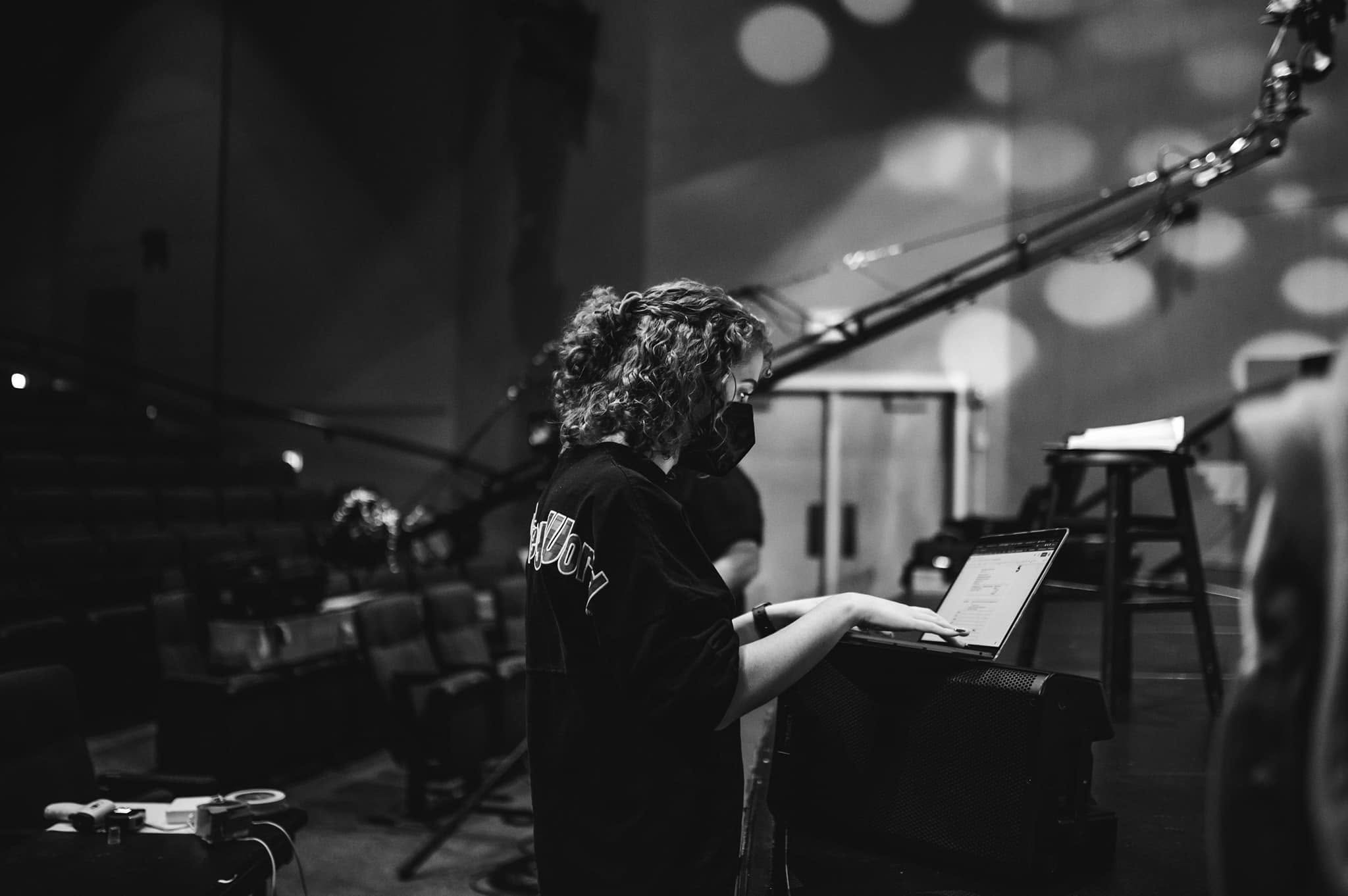Person with curly hair working on a laptop in a dimly lit room with stage lights and equipment.