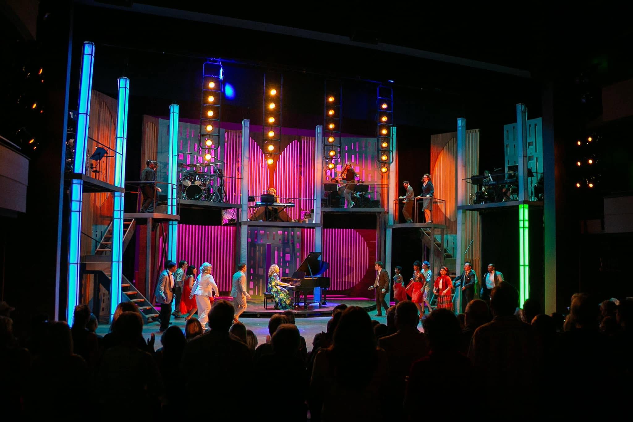 Stage with performers, colorful lighting, and audience in foreground, set for a musical or theatrical performance.