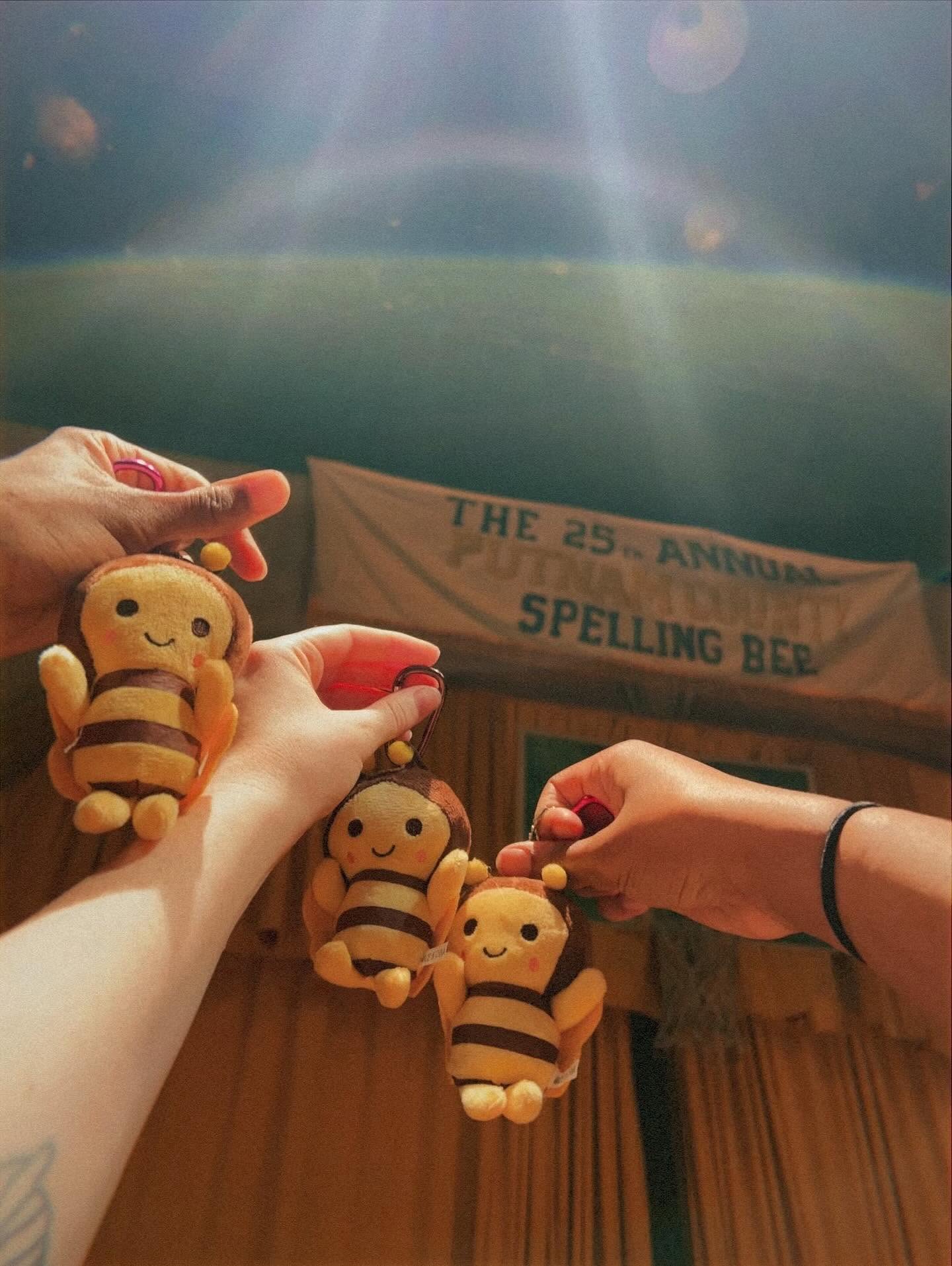 People hold bee plush toys in front of a banner and a window with sunlight, indoors. The banner reads 'The 25th Annual Spelling Bee.'