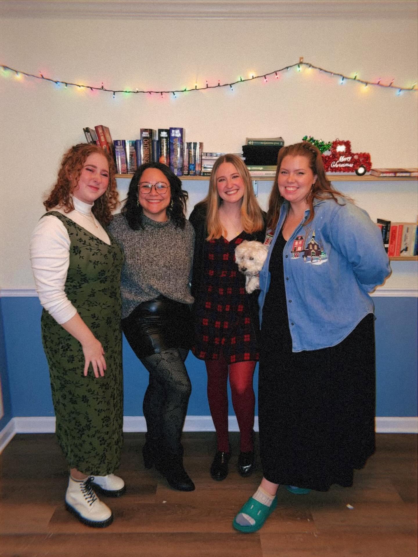 Four women standing together indoors, smiling, with books and colorful string lights above them.