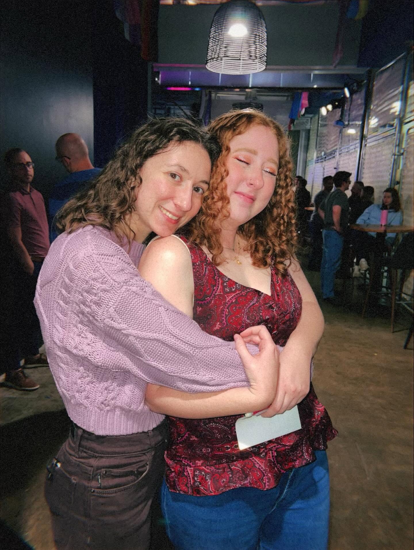Two women hugging and smiling in a social setting with people and a bar in the background.