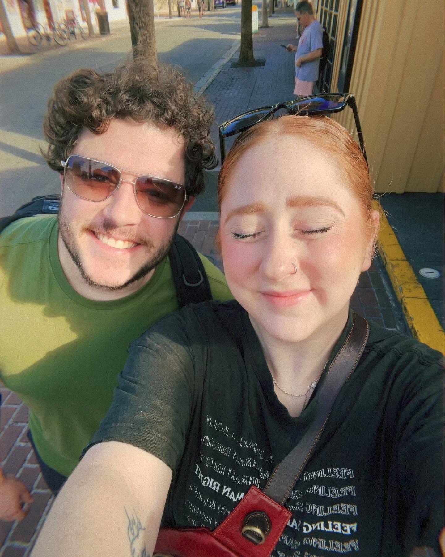 Two people taking a selfie outdoors, smiling with trees and a person in the background.