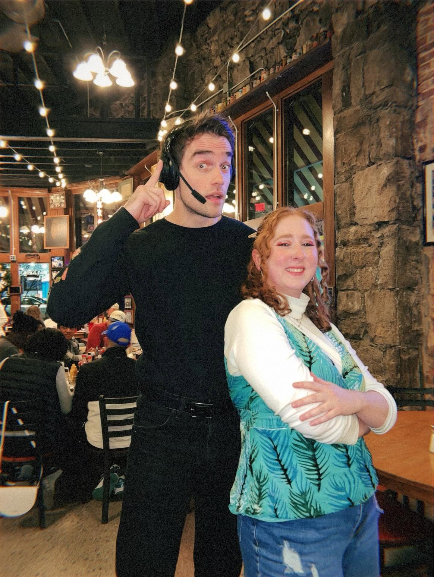 A young man wearing a headset and black shirt stands next to a smiling woman in a white and green patterned top inside a restaurant with stone walls and string lights.