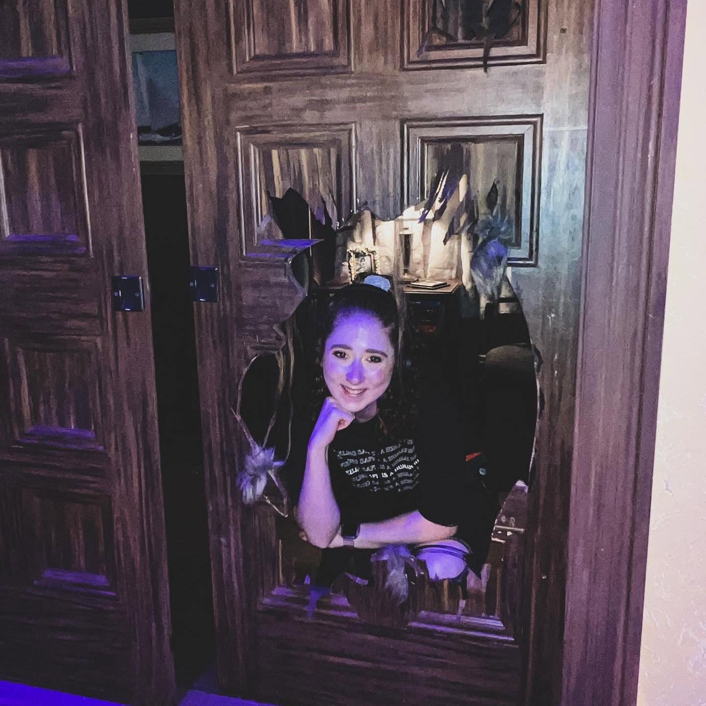 Person with long hair and makeup, sitting with chin on hand, reflected in a mirror with wooden panels, dim lighting.