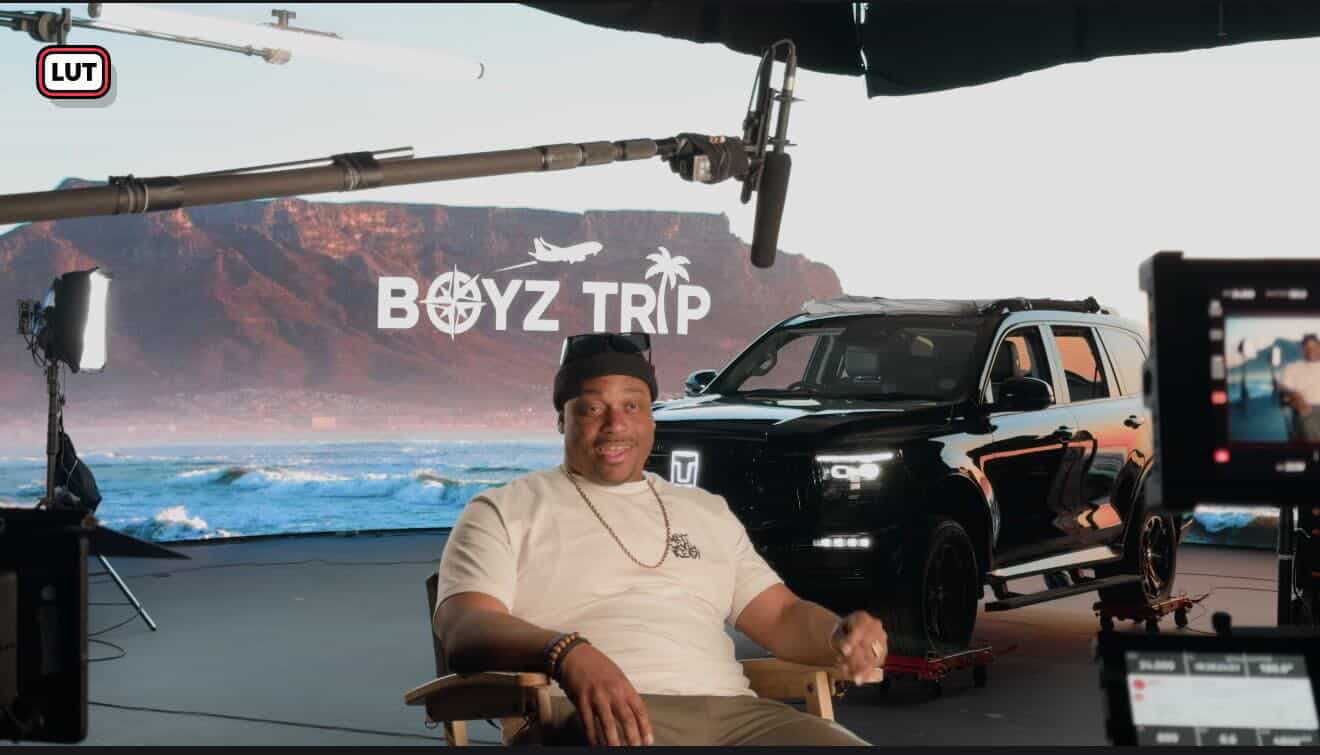 Person sitting in front of a backdrop with a boat and mountains, with a black vehicle and camera equipment nearby.
