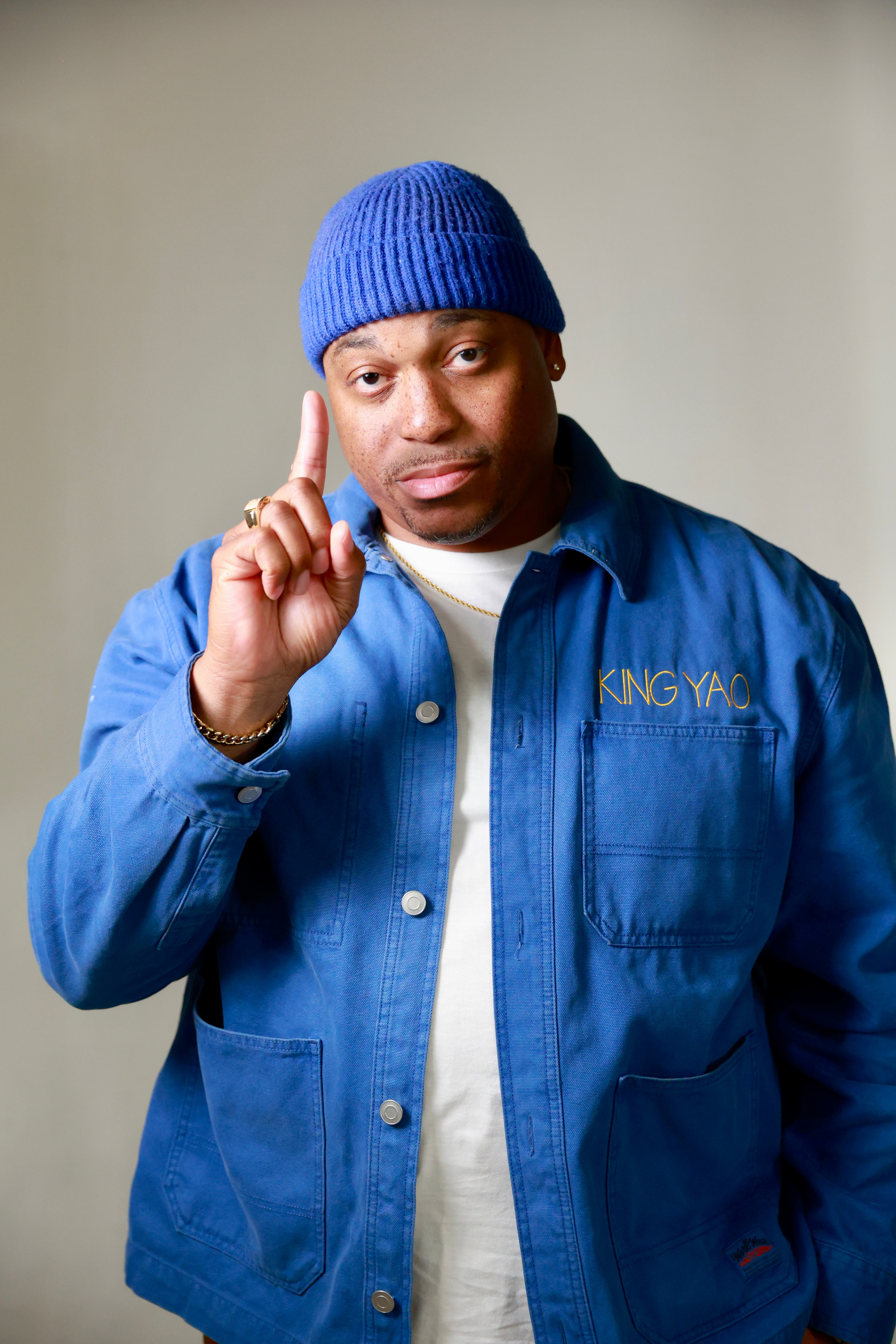 Man wearing a blue beanie and jacket, holding finger to his temple, against a plain background.