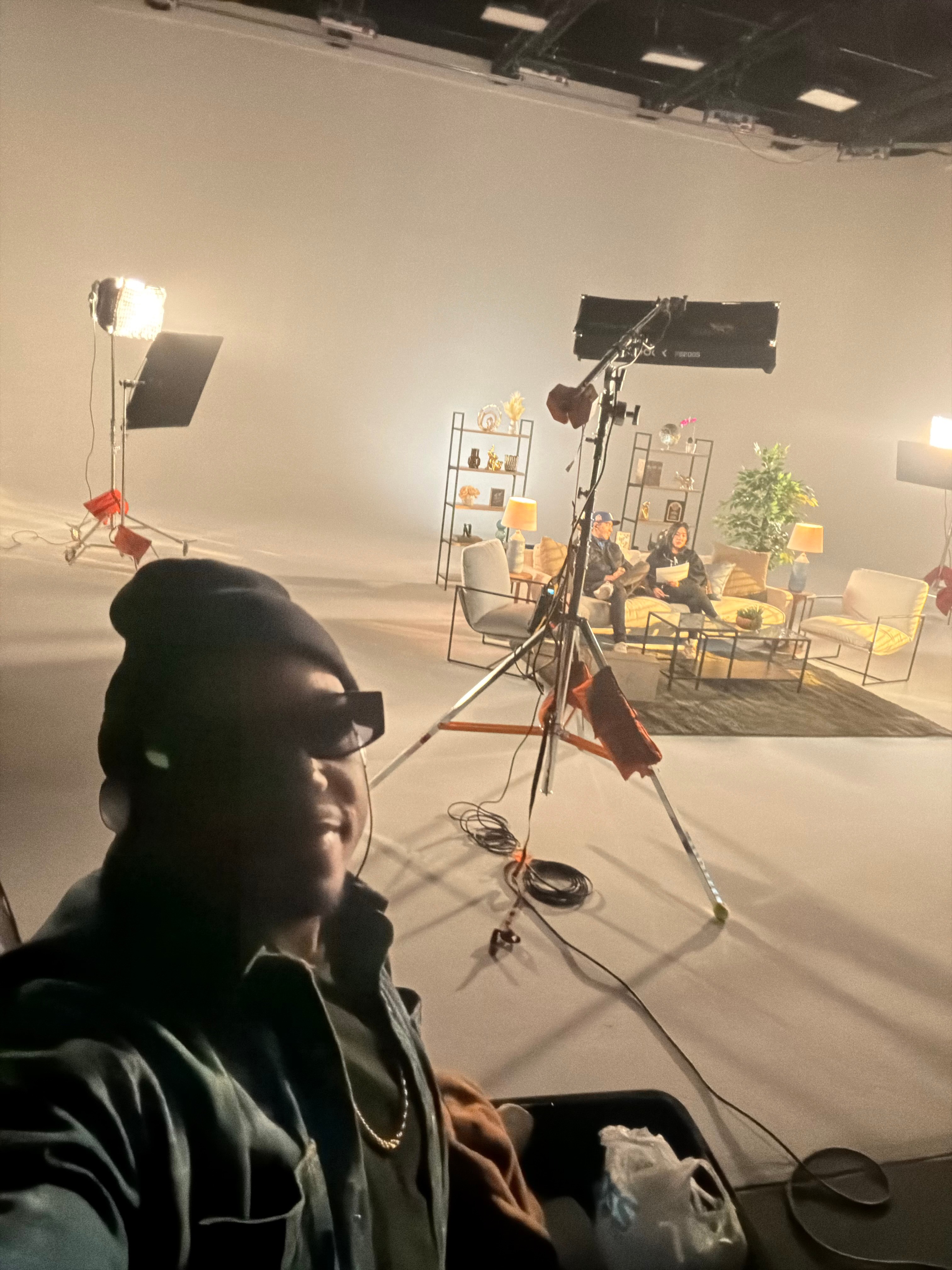 Person taking selfie in a studio with lighting, camera equipment, and a seating area with chairs and plants.
