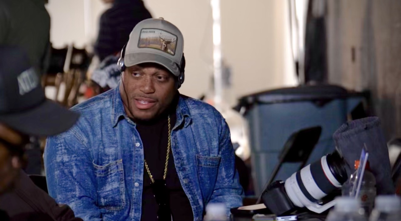 Young man wearing a baseball cap and denim jacket sitting indoors, with camera equipment nearby.