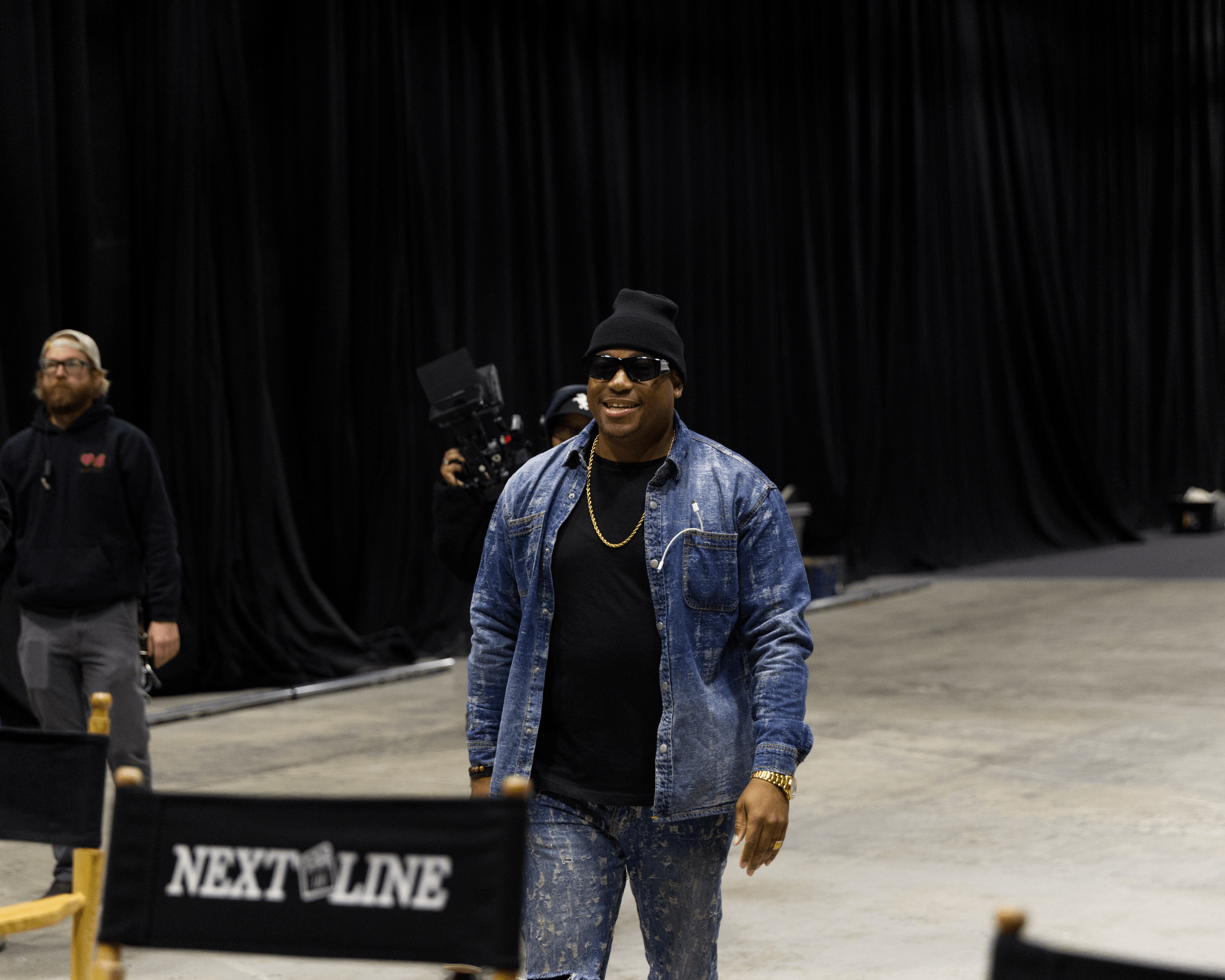 Person wearing sunglasses, black beanie, denim jacket, and black shirt walking indoors with black curtains in background.