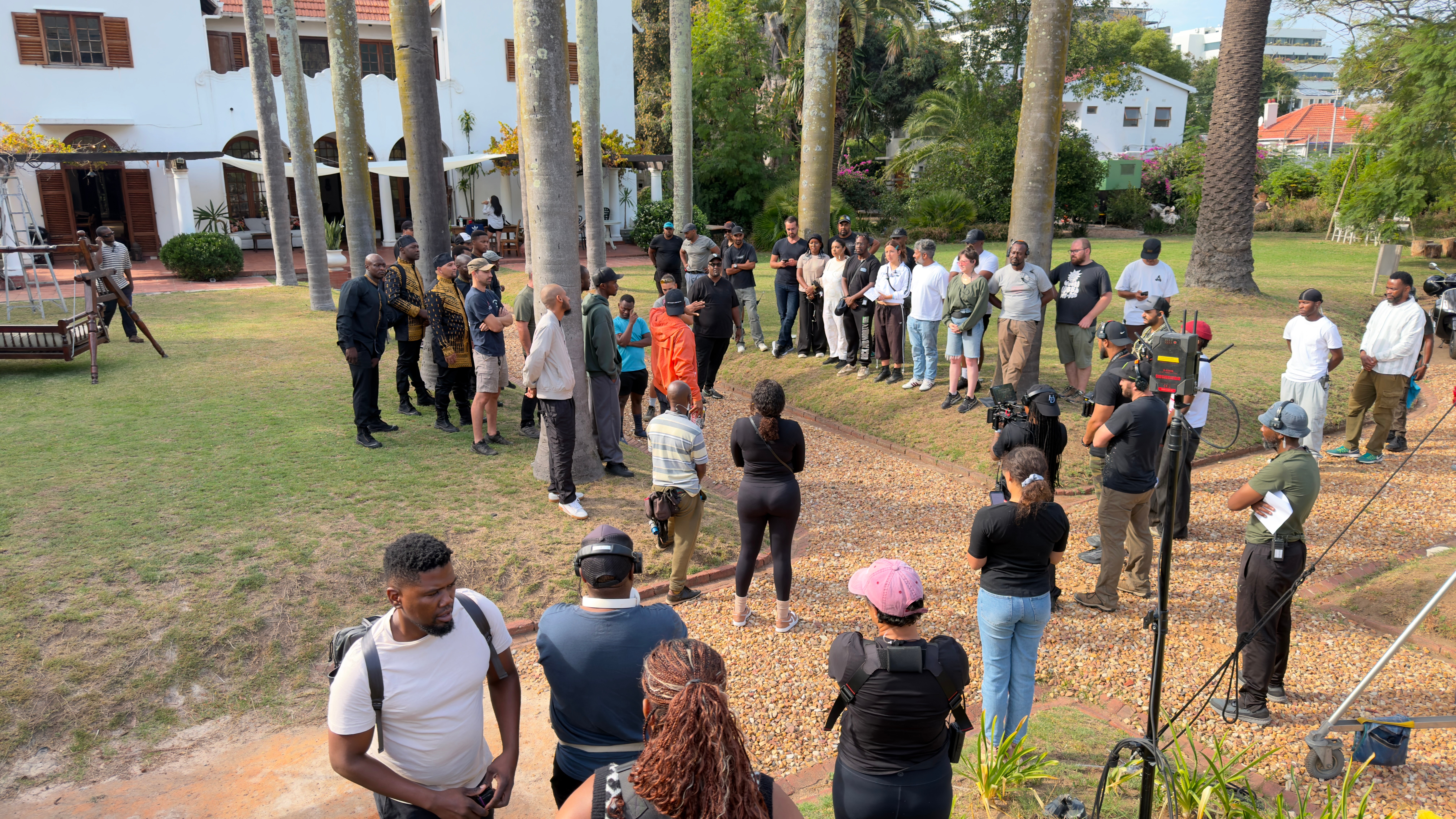 Group of people gathered outdoors in a park-like setting with trees and buildings in the background.