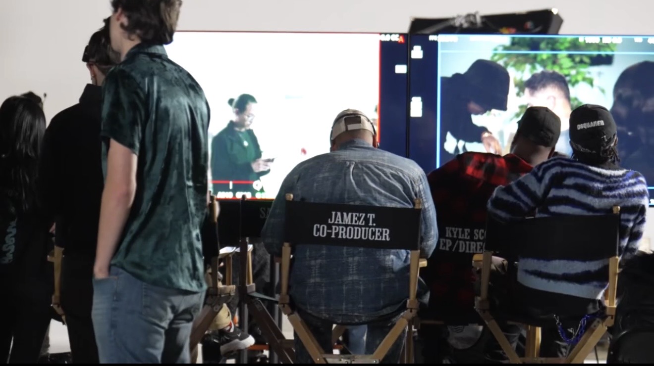 People watching a film or video on a large screen, seated in chairs, with some standing nearby, in a dark room.
