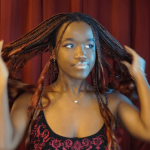 Young woman with braided hair adjusting her hair, standing in front of red curtains, wearing a black and red top, looking to the side.