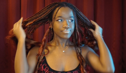 Young woman with braided hair adjusting her hair, standing in front of red curtains, wearing a black and red top, looking to the side.