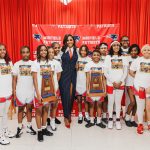 Group of young female athletes in white shirts and red shorts with a woman in a suit, standing in front of red curtains.