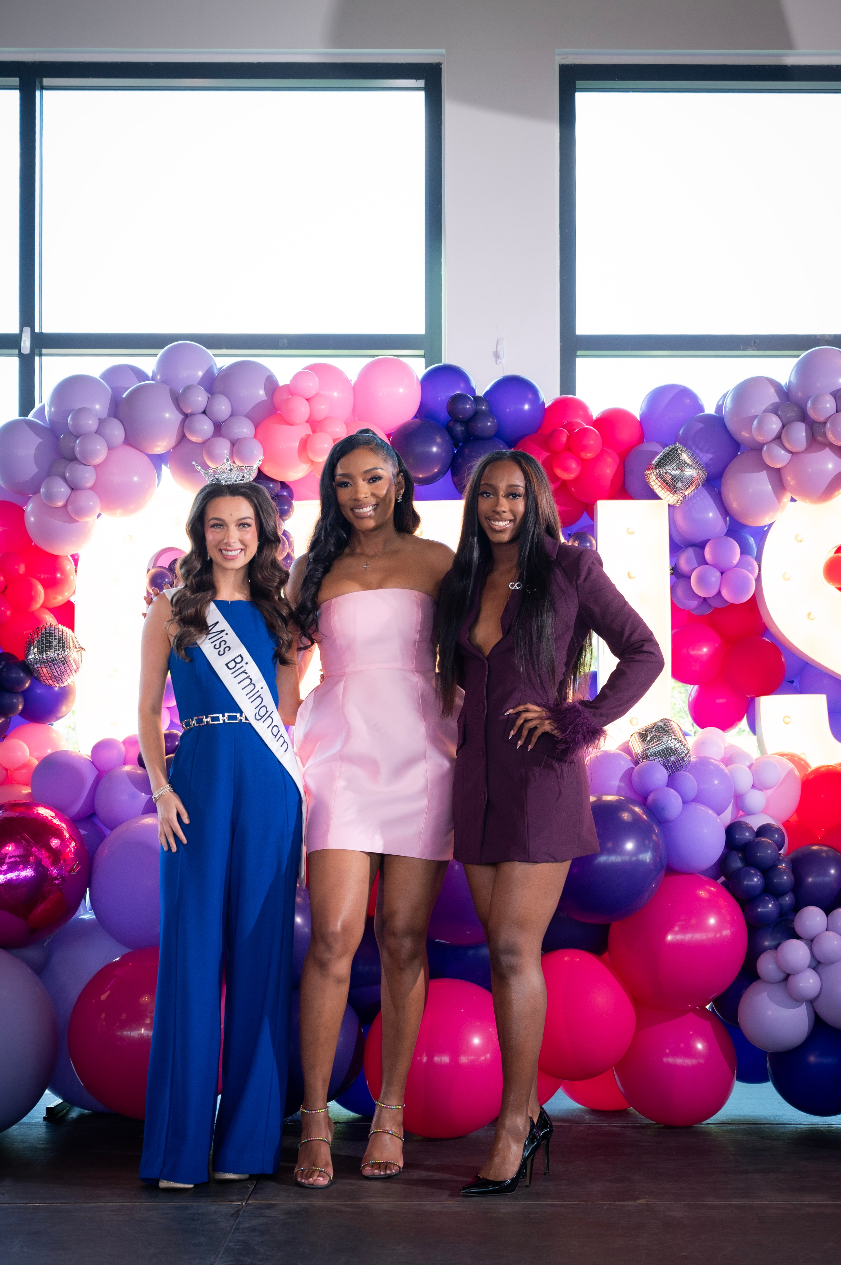 Three women stand in front of a colorful balloon backdrop, smiling, with windows in the background.