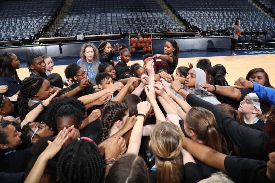 Group of young girls in sportswear forming a circle with hands joined in the center inside a stadium.